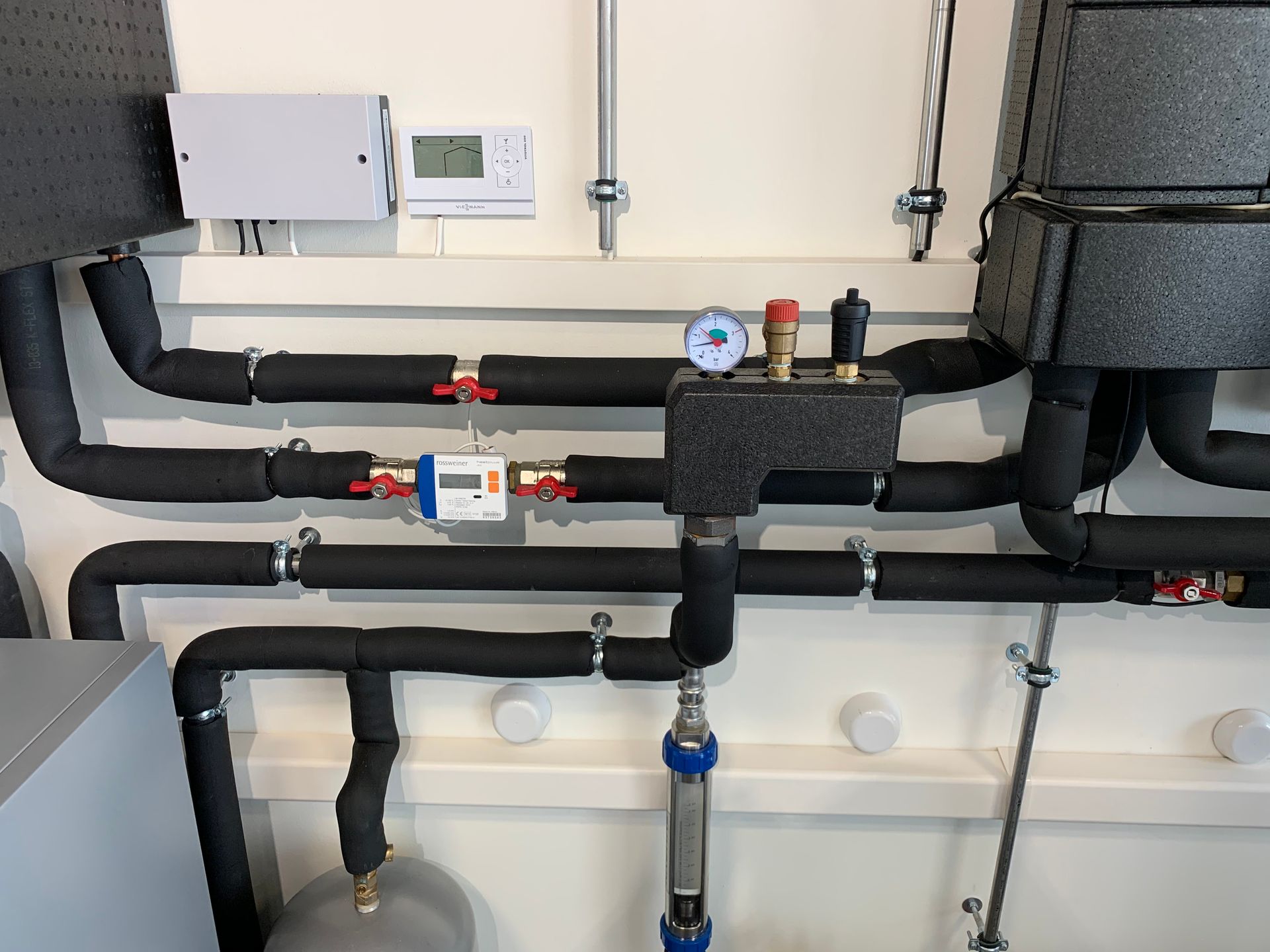 Tuyaux avec isolation et vannes, reliés à un boîtier de commande de système de chauffage et à un thermostat sur un mur blanc.