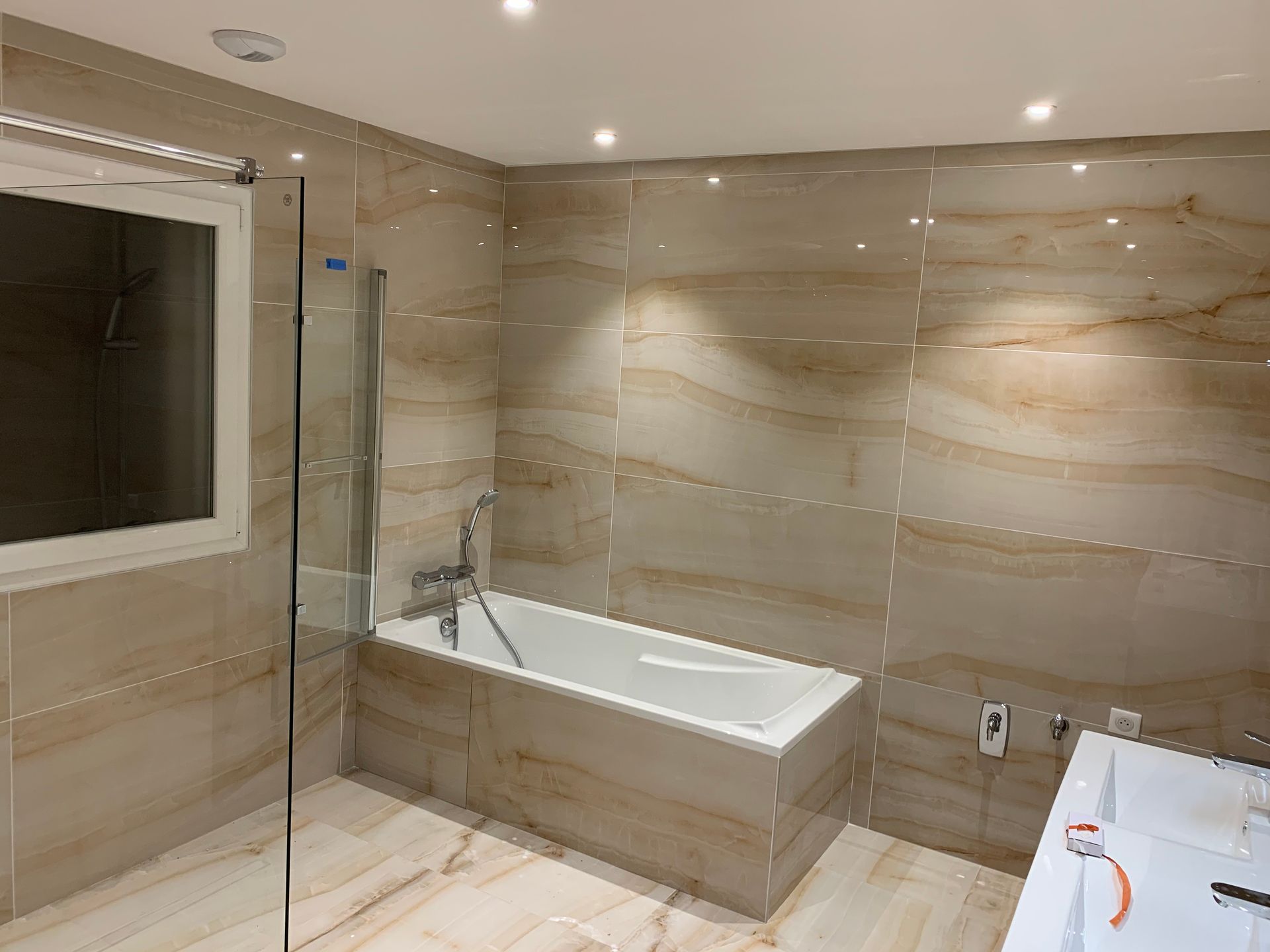 Salle de bains avec carrelage effet marbre beige, baignoire blanche et fenêtre. Paroi de douche en verre.