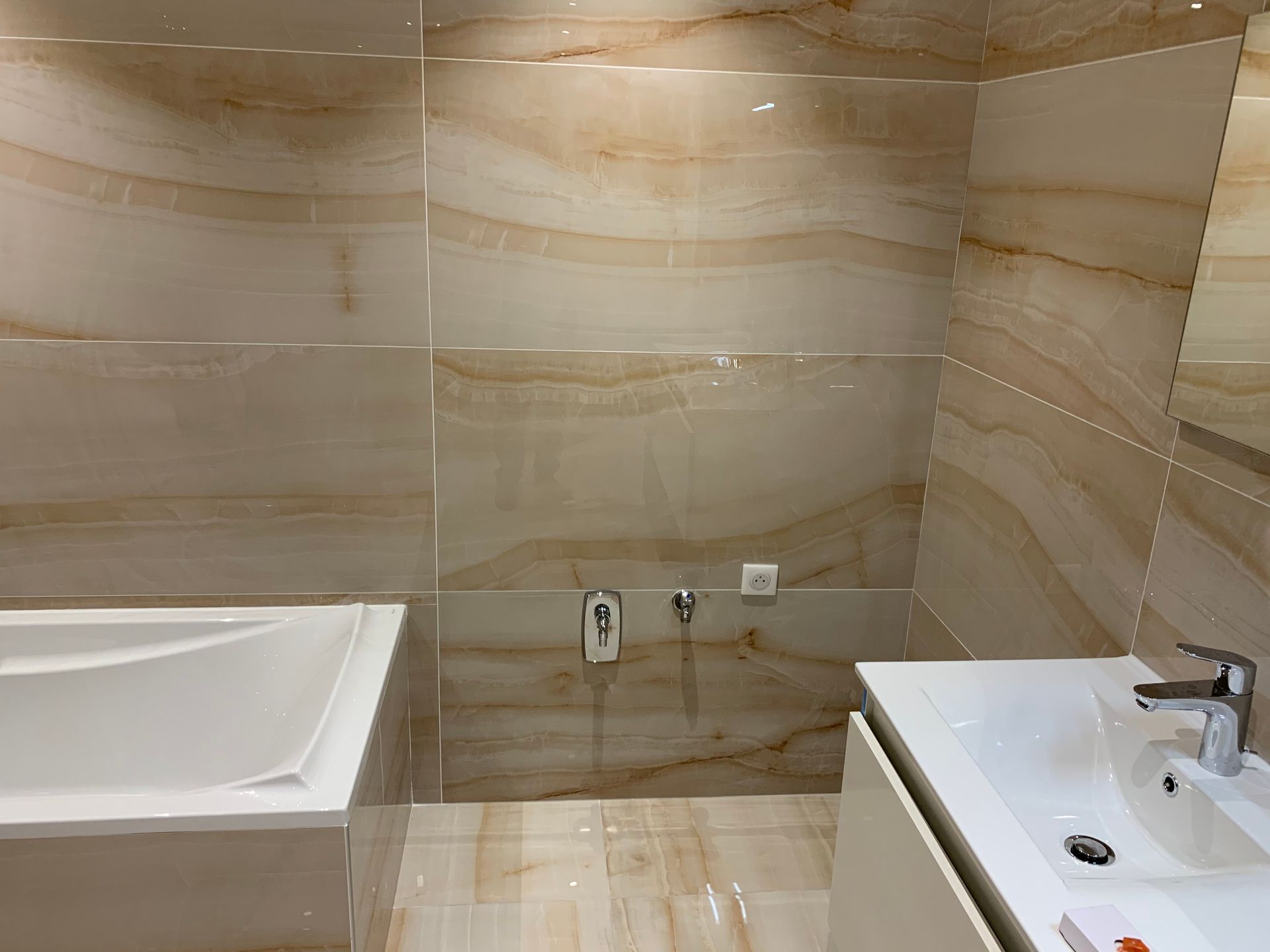 Salle de bains avec carrelage beige effet marbre, baignoire blanche, lavabo et accessoires.