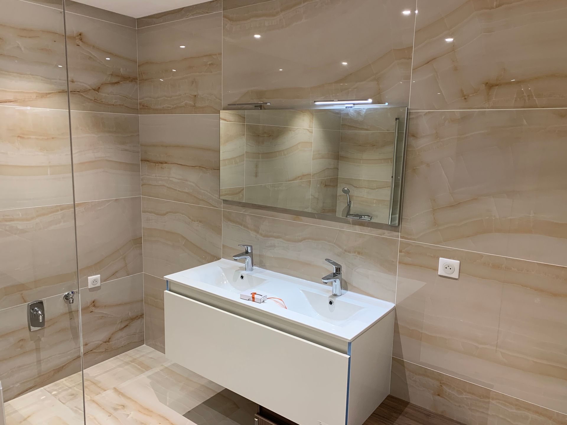 Salle de bains avec carrelage imitation marbre beige. Meuble-lavabo double blanc avec miroir, robinetterie chromée et douche en verre.