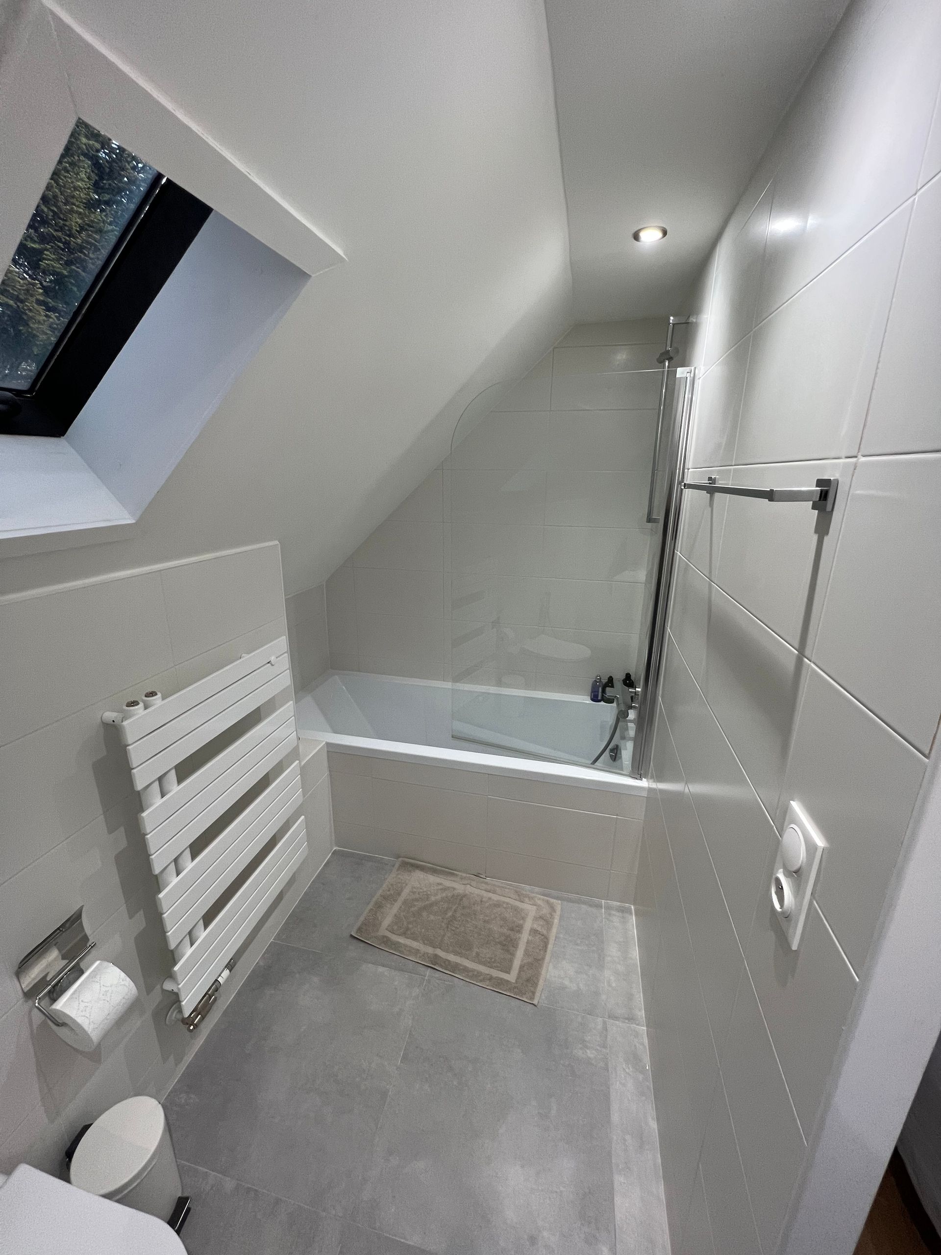 Petite salle de bains avec plafond mansardé, baignoire et porte-serviettes ; tons blancs et gris.