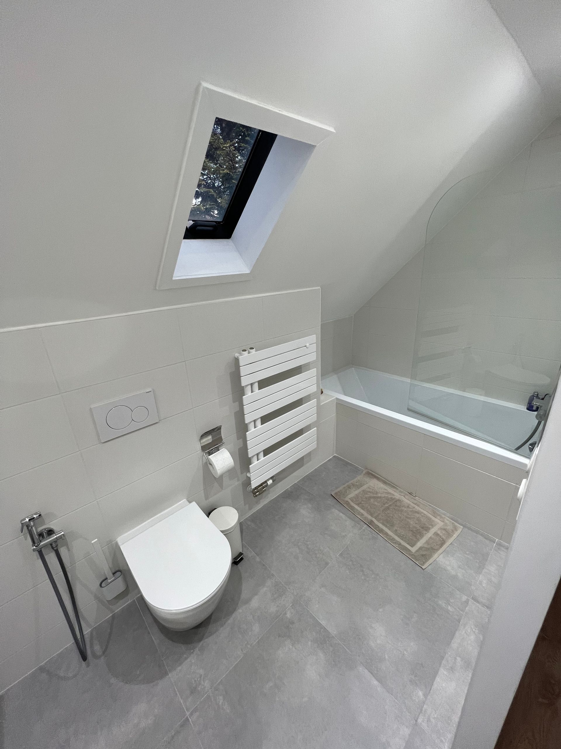 Salle de bains avec plafond mansardé, puits de lumière, toilettes, baignoire et sèche-serviettes. Carrelage gris au sol, murs et sanitaires blancs.