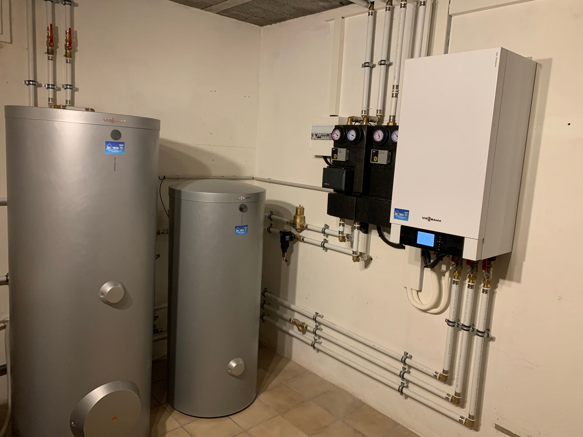 Réservoirs de chauffe-eau argentés et chaudière blanche dans une buanderie avec tuyaux et panneaux de commande.