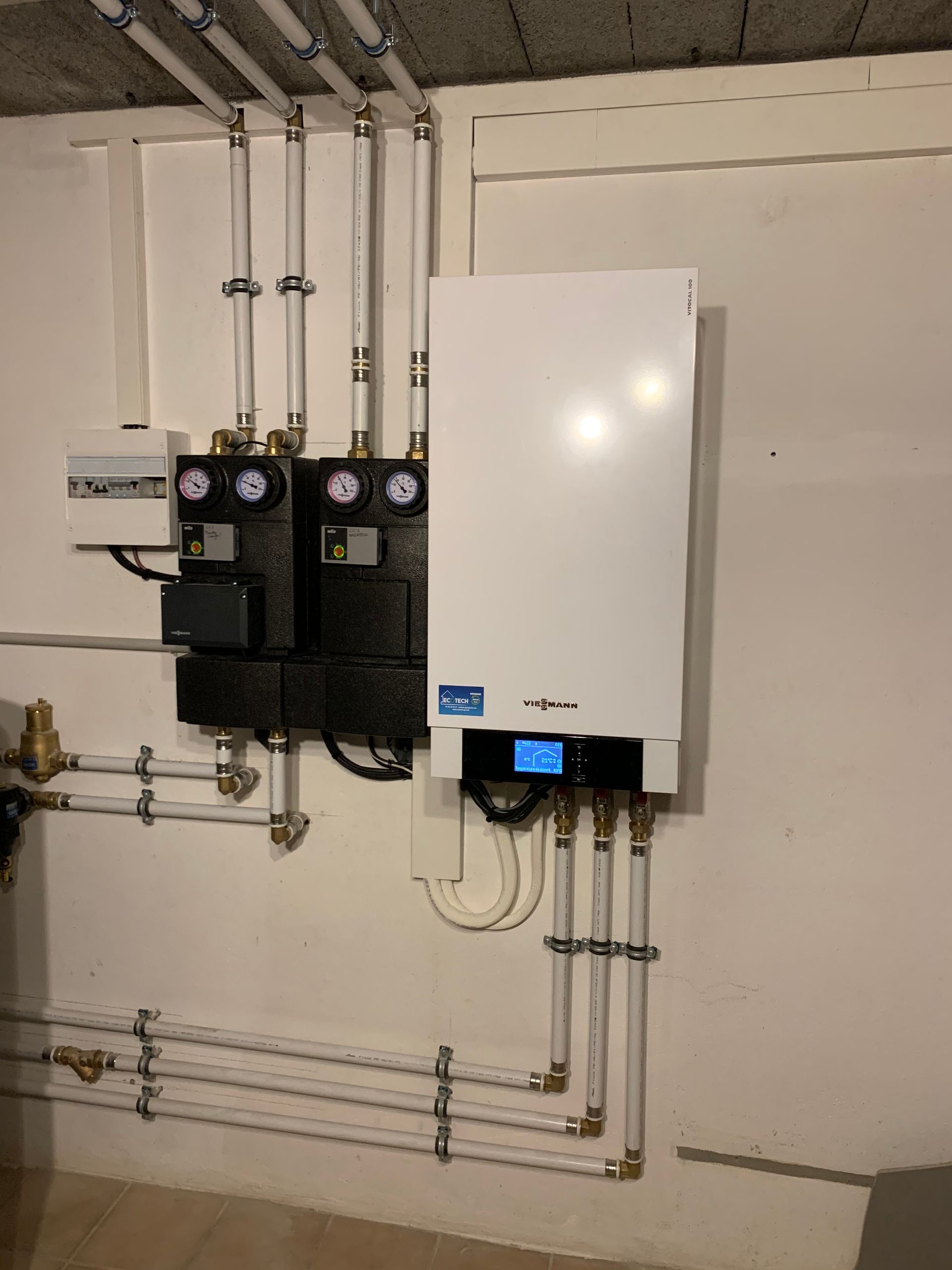 Composants de système de chauffage blancs et noirs montés sur un mur blanc avec tuyauterie visible.