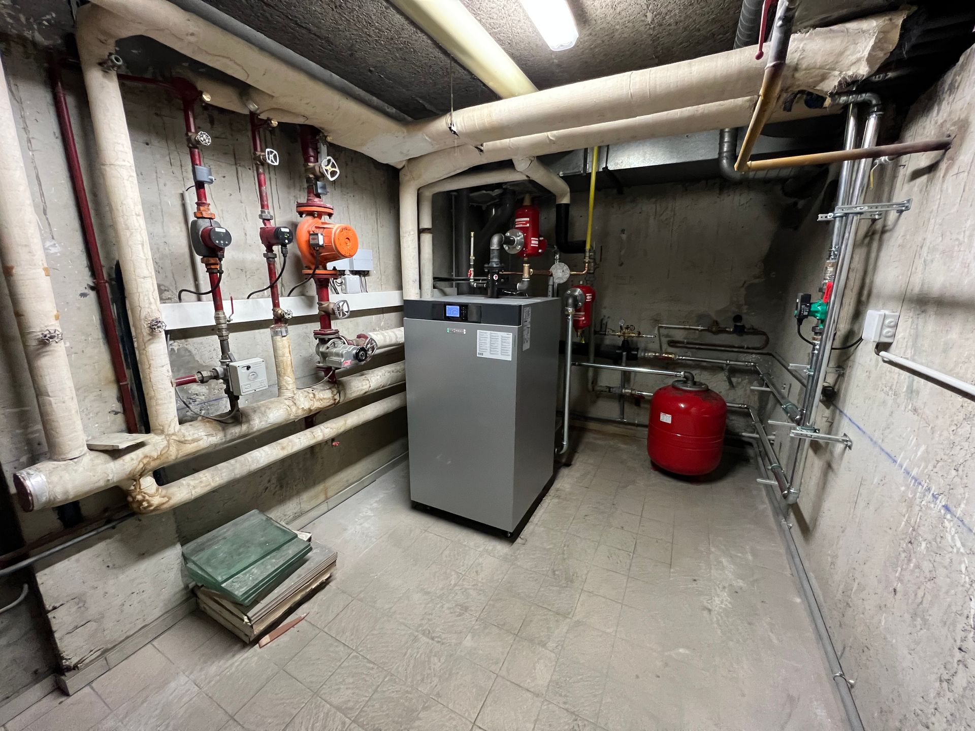 Local de chauffage avec tuyaux, chaudière grise, réservoir rouge et vannes contre murs en béton.