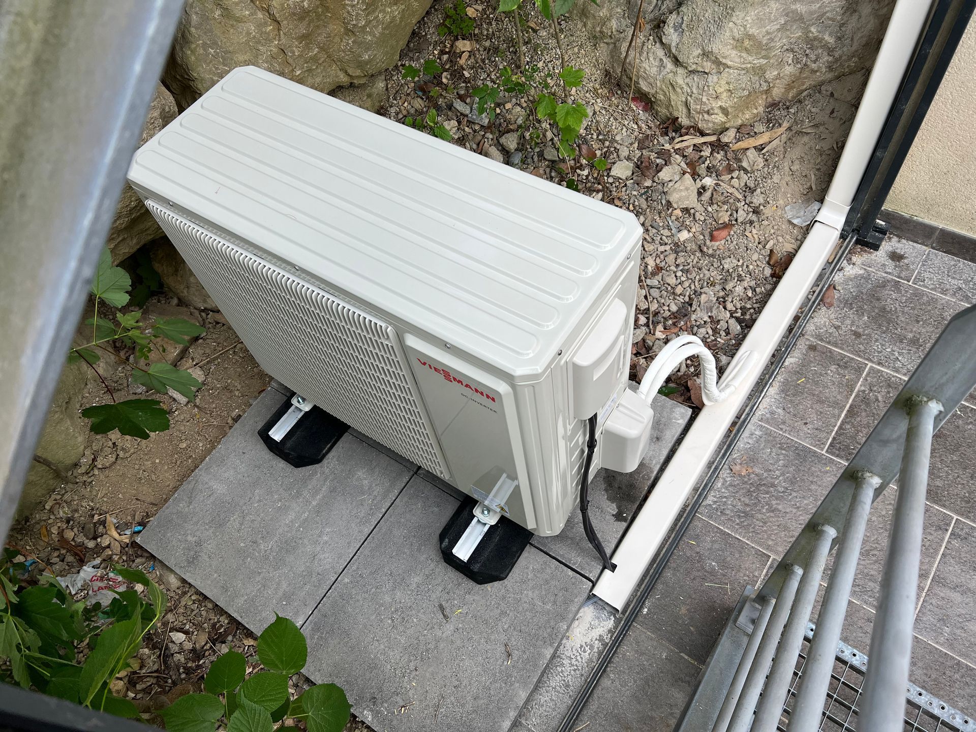 Une unité de pompe à chaleur blanc cassé sur des blocs de béton, à côté d'un tuyau d'évacuation et d'escaliers, à l'extérieur.