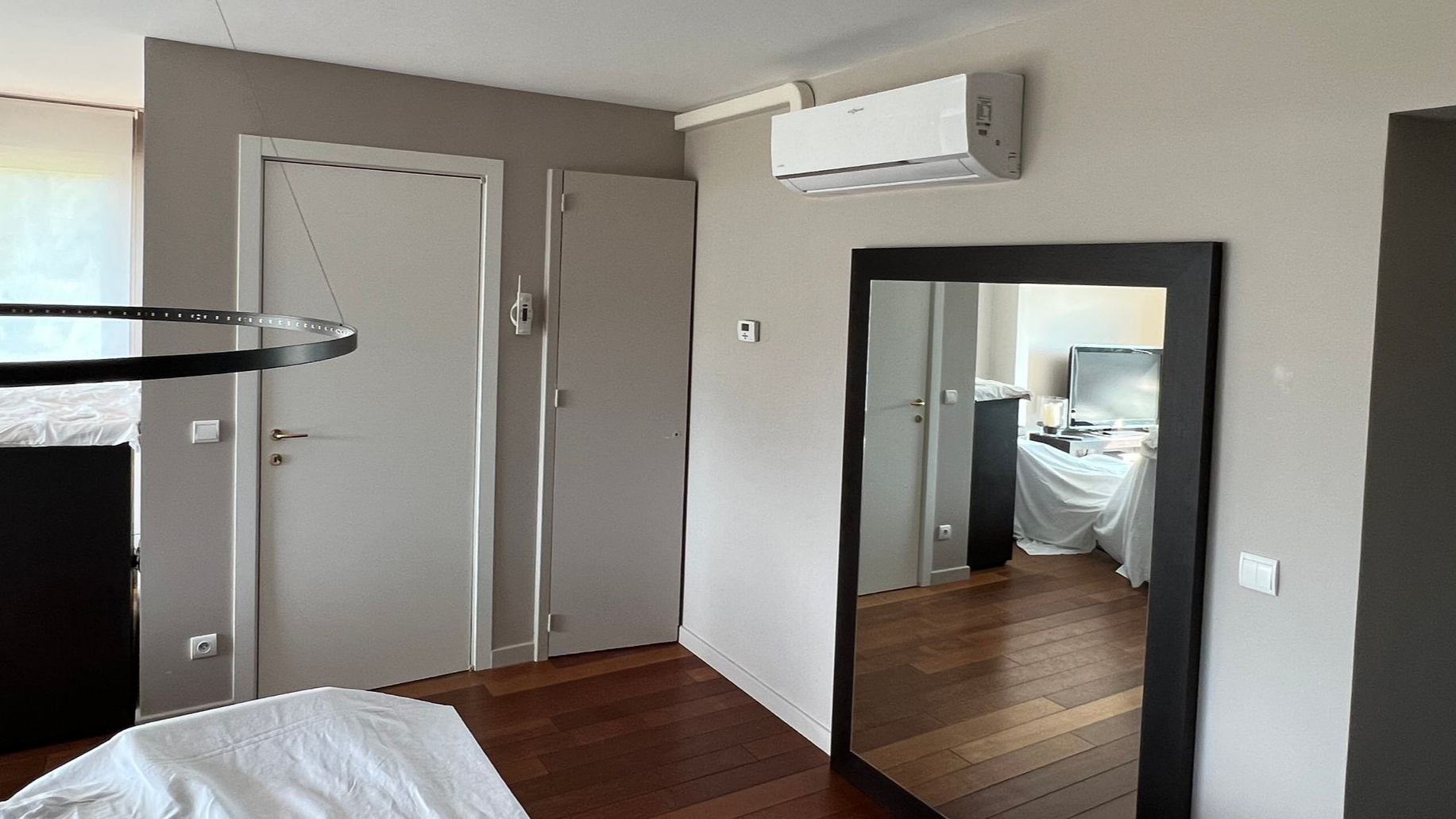 Chambre avec un grand miroir reflétant une pièce avec parquet. Portes et climatiseur visibles.