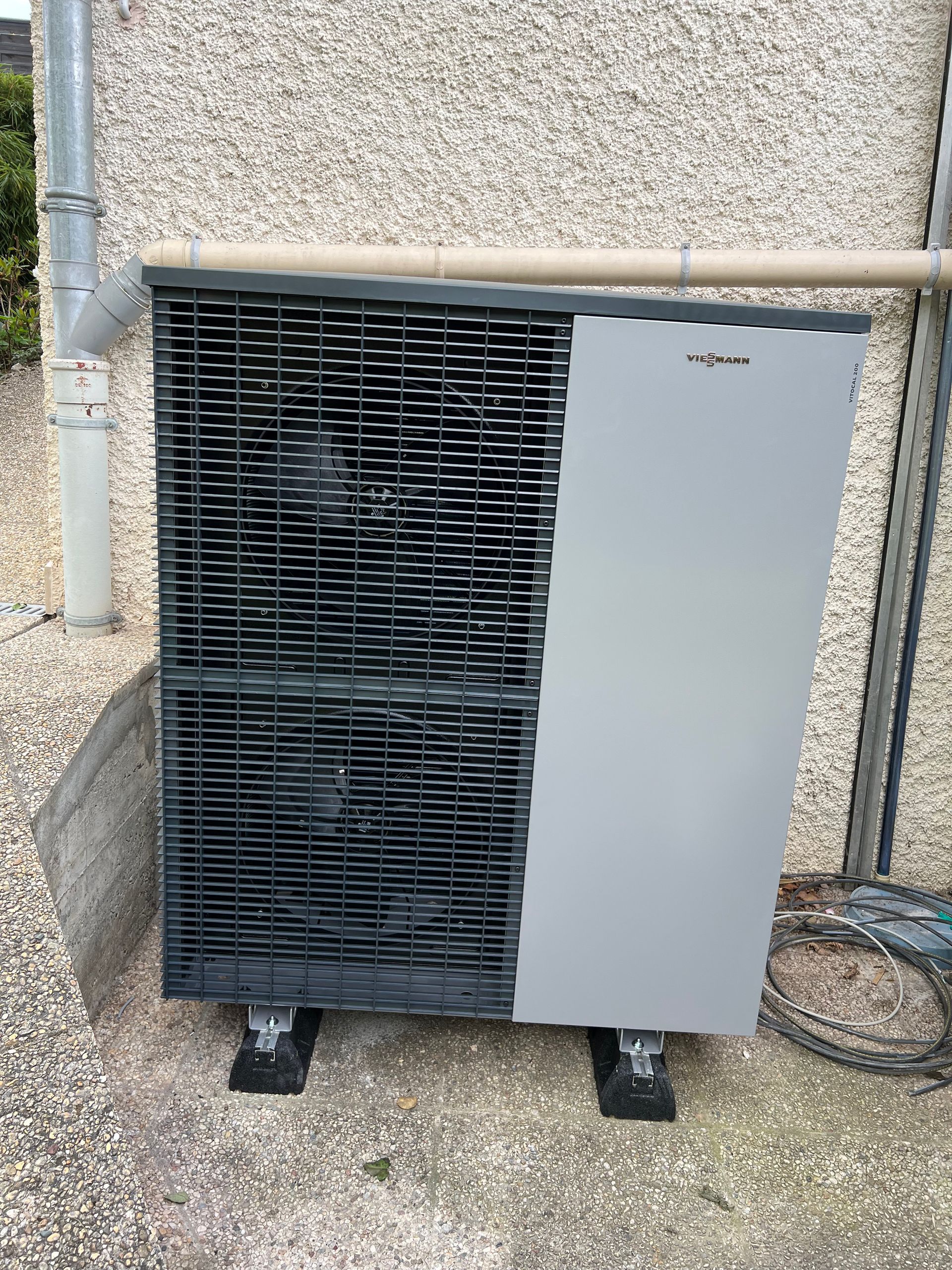 Pompe à chaleur montée sur blocs contre un mur texturé, avec une grille métallique et un panneau gris.