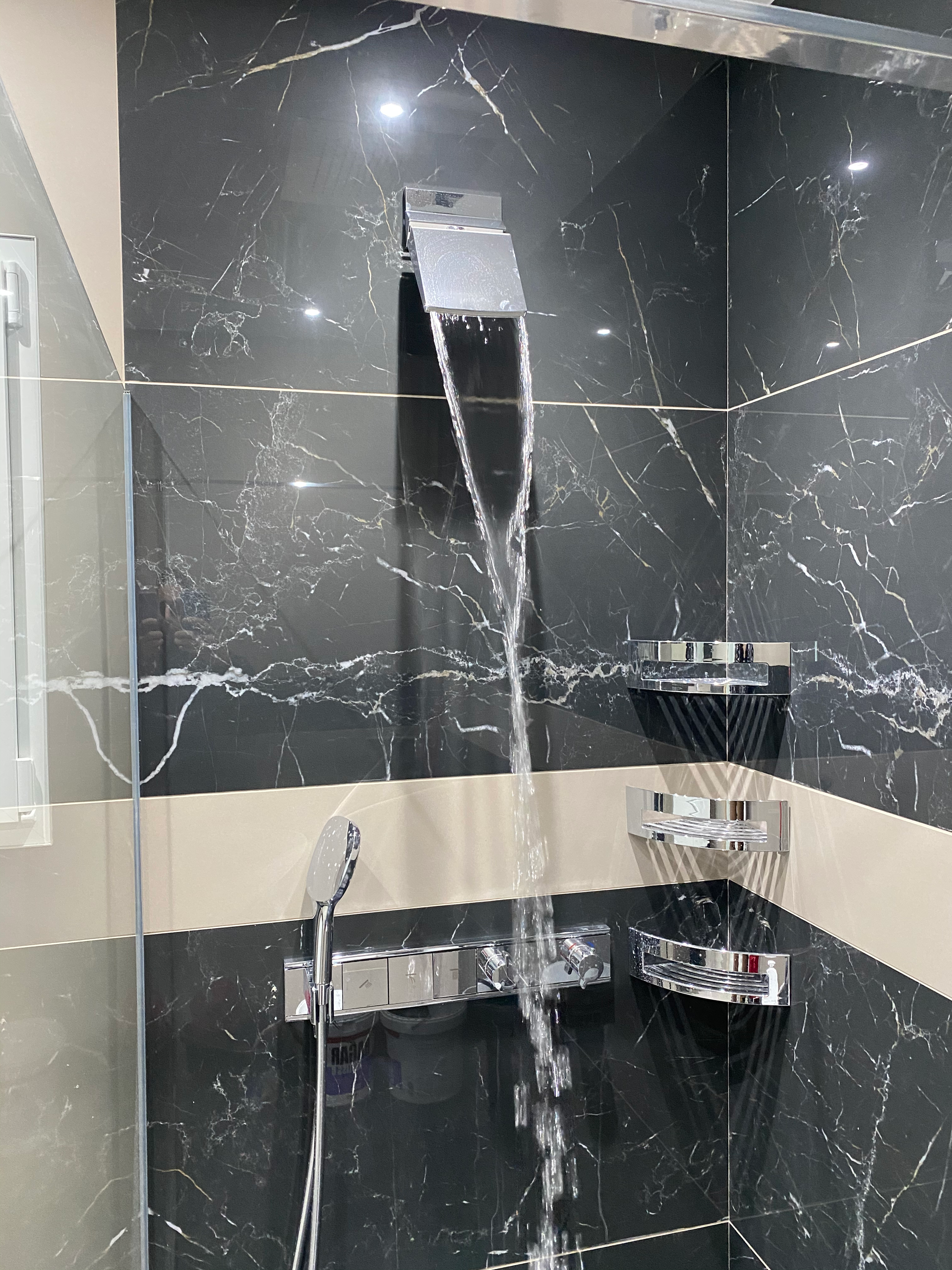Douche avec robinet en cascade ; l'eau coule sur des carreaux de marbre noir et des étagères avec des articles de toilette.