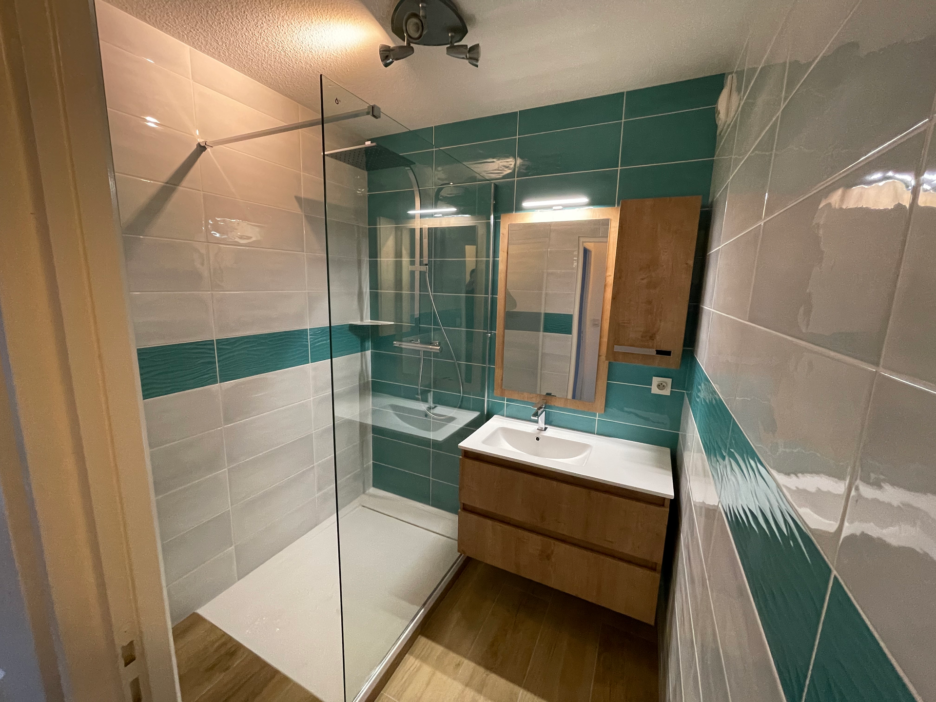 Petite salle de bains avec des murs carrelés bleu sarcelle et gris, une douche en verre, une vanité en bois et un miroir.
