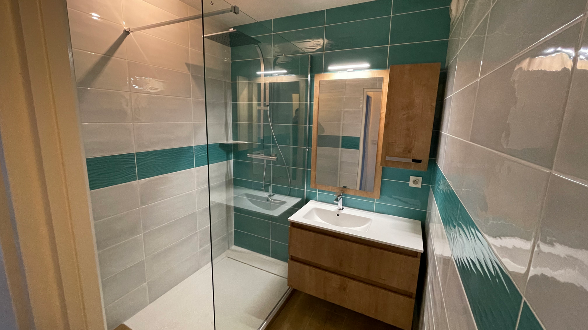 Petite salle de bains avec des murs carrelés bleu sarcelle et gris, une douche en verre, une vanité en bois et un miroir.