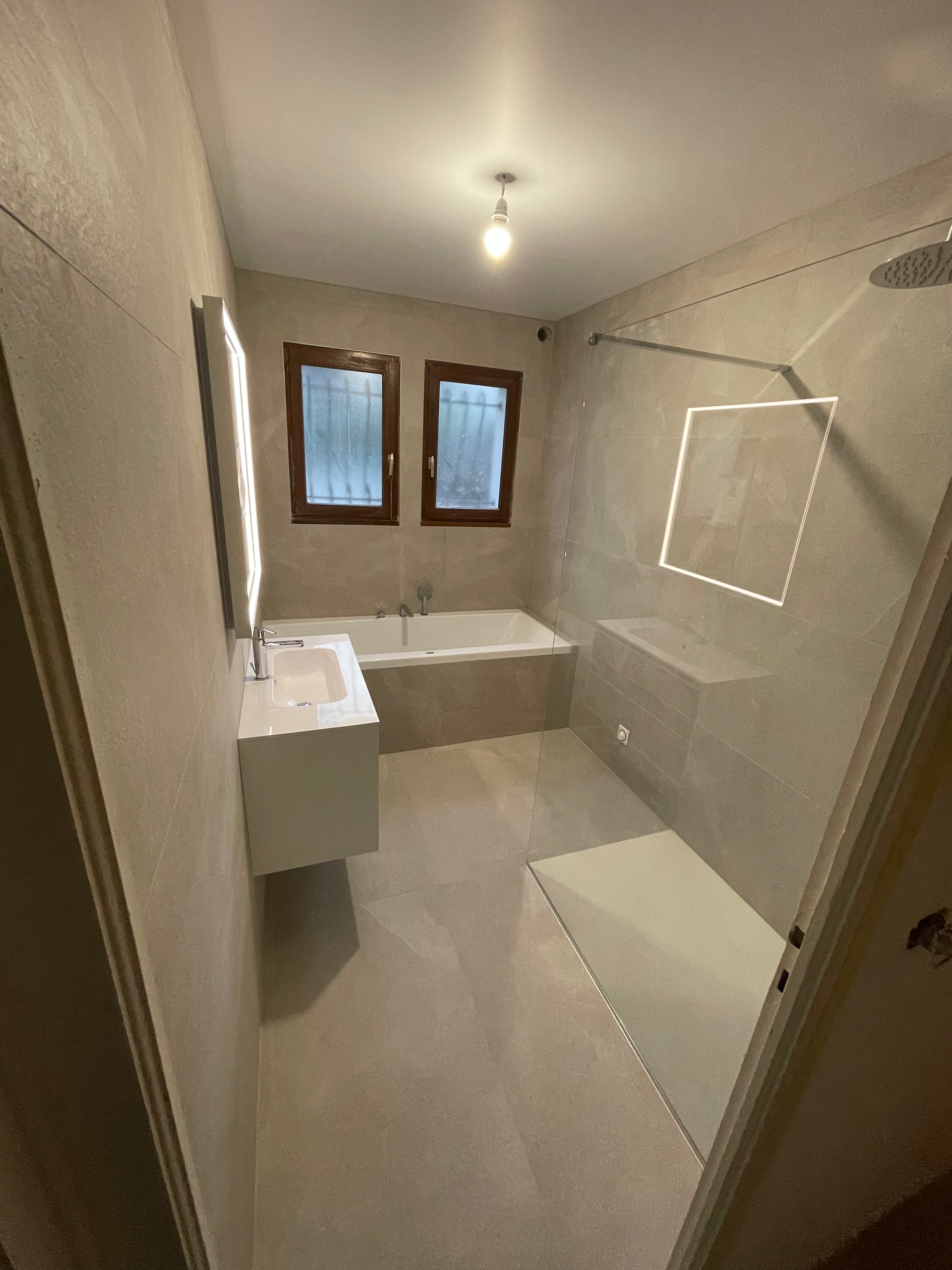 Salle de bains avec baignoire, douche en verre, deux fenêtres et une vanité flottante.