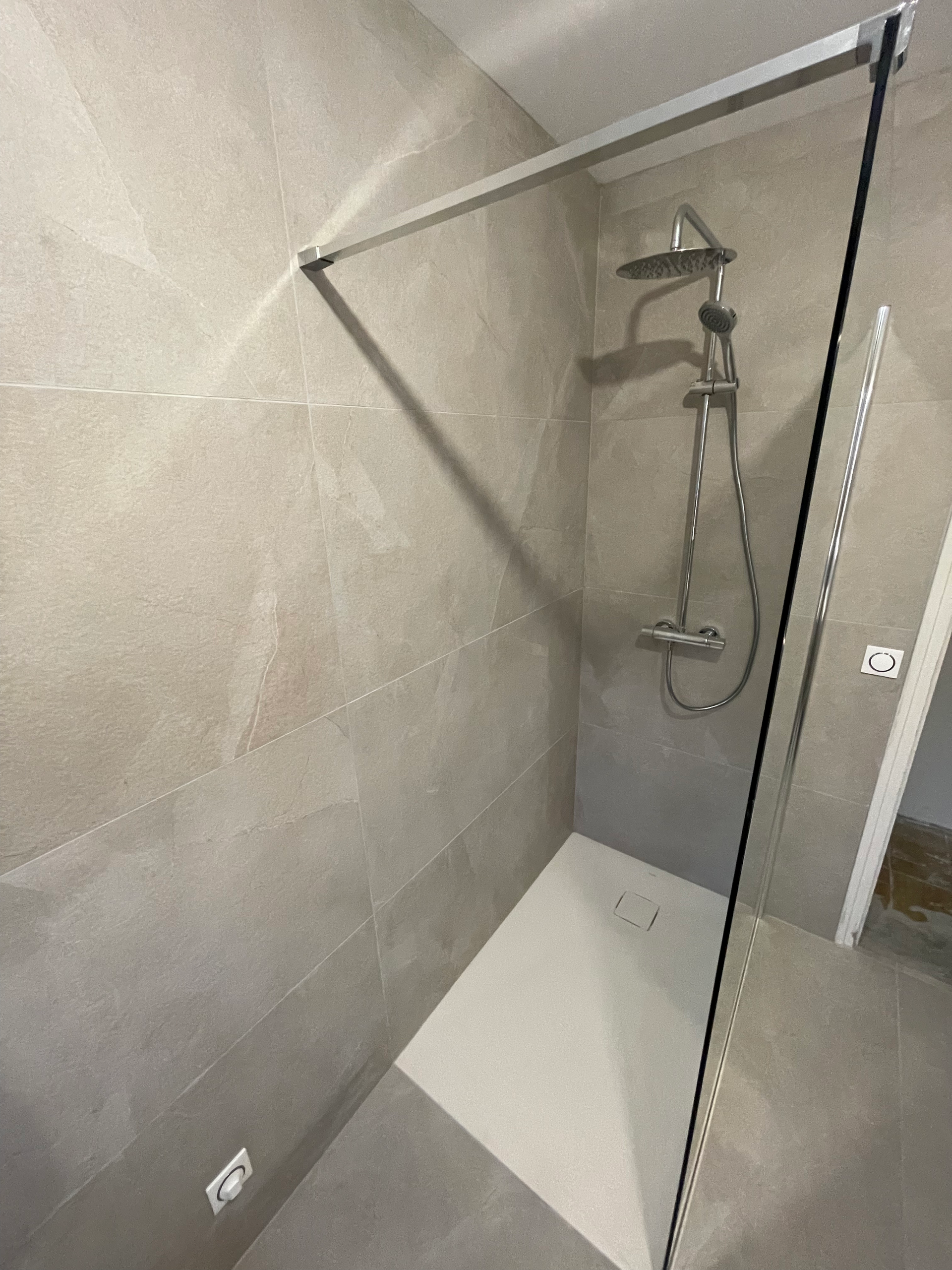 Cabine de douche avec murs carrelés gris, porte vitrée et accessoires de douche argentés.