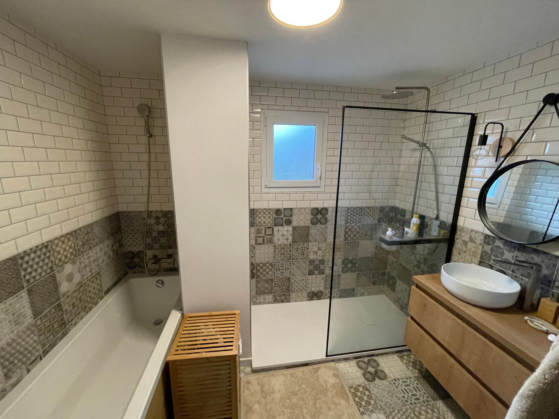 Salle de bains moderne avec baignoire, douche, vanité en bois, carrelage à motifs et miroir rond.
