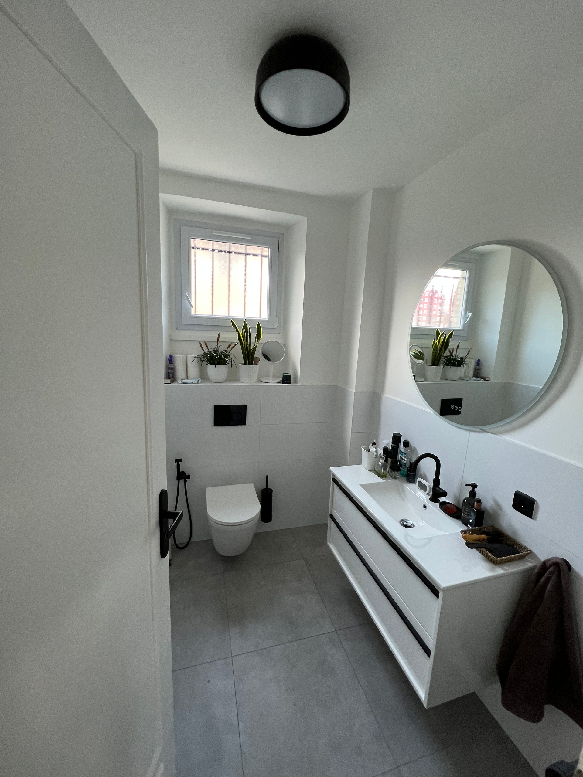 Salle de bains blanche moderne avec sol gris, vanité blanche, miroir rond et accents noirs.