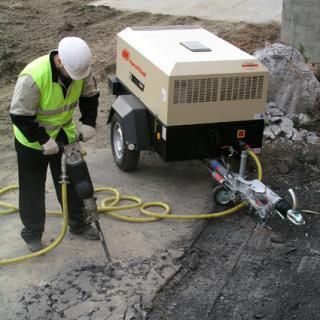 Obrero de la construcción operando un martillo neumático sobre asfalto, conectado a un compresor de aire montado en un remolque.