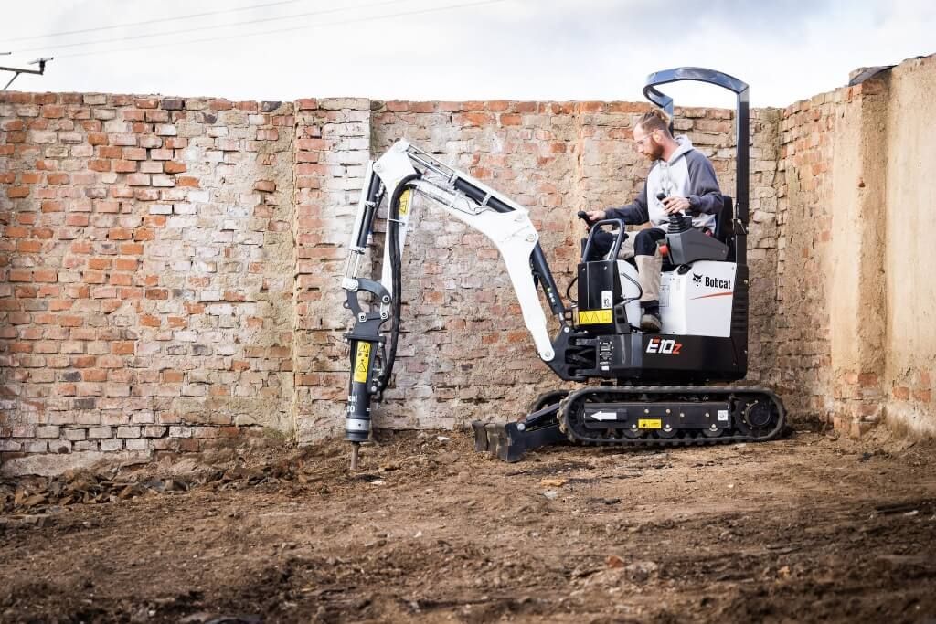 Un hombre conduce una pequeña excavadora frente a una pared de ladrillos.