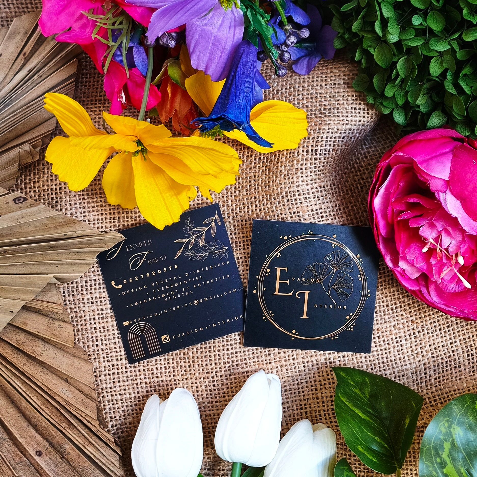 Deux cartes de visite carrées noires à dorure à chaud reposent sur une surface en toile de jute texturée, entourées de fleurs colorées.