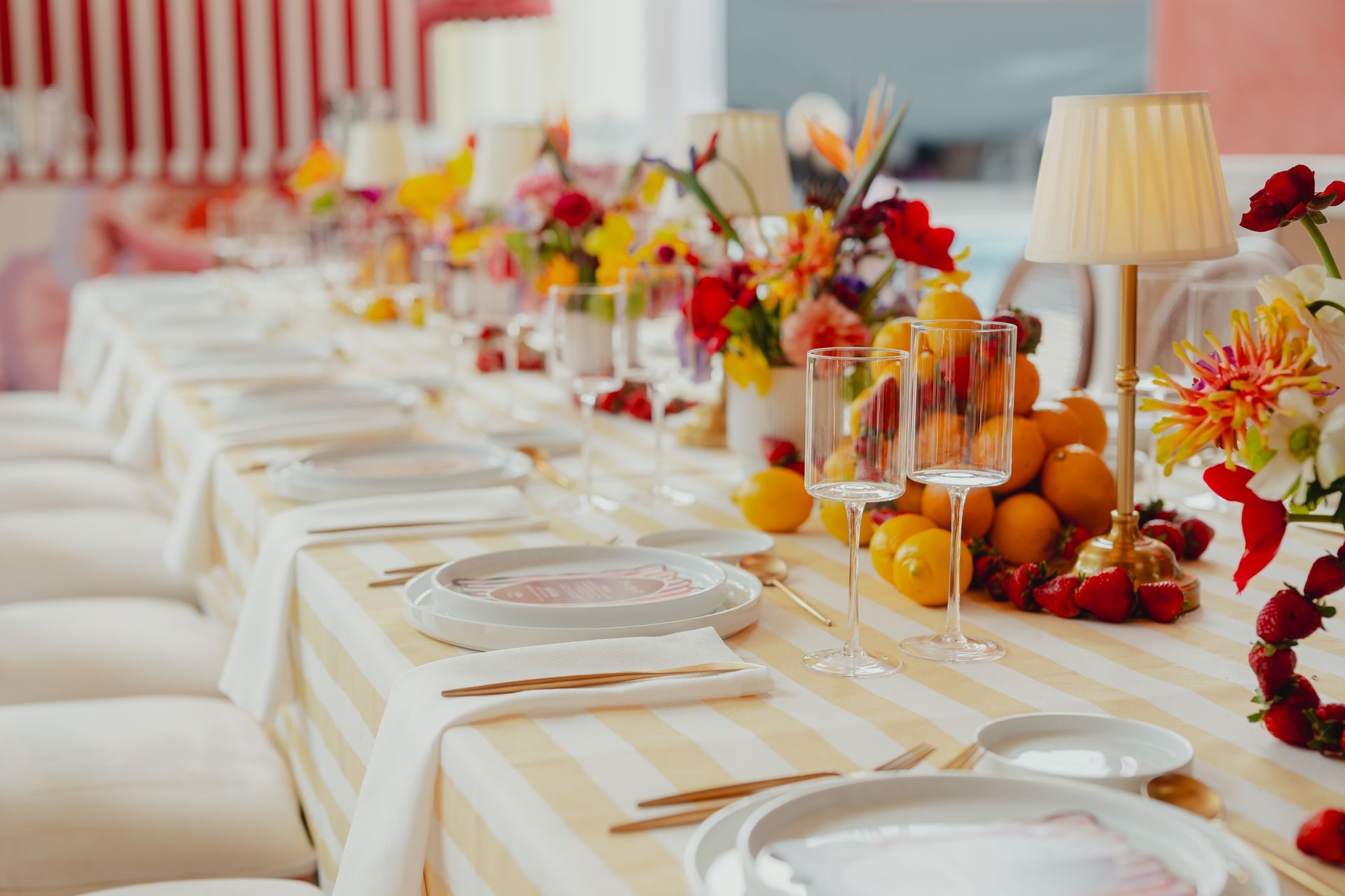 Longue table à manger dressée pour un événement avec du linge de table à rayures blanches et jaunes, des centres de table floraux, des citrons et des petites lampes.