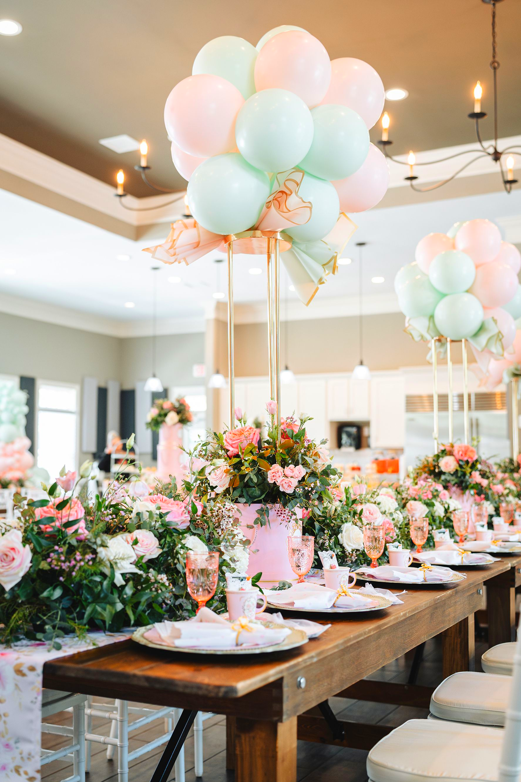 De longues tables ornées de centres de table floraux rose poudré et menthe, surmontés de grappes de ballons assorties, dans une pièce lumineuse.