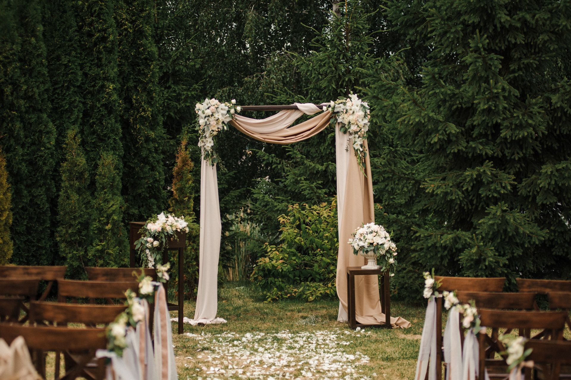 Un décor de cérémonie de mariage en plein air avec une arche drapée, des compositions florales et des chaises en bois dans un jardin verdoyant luxuriant.