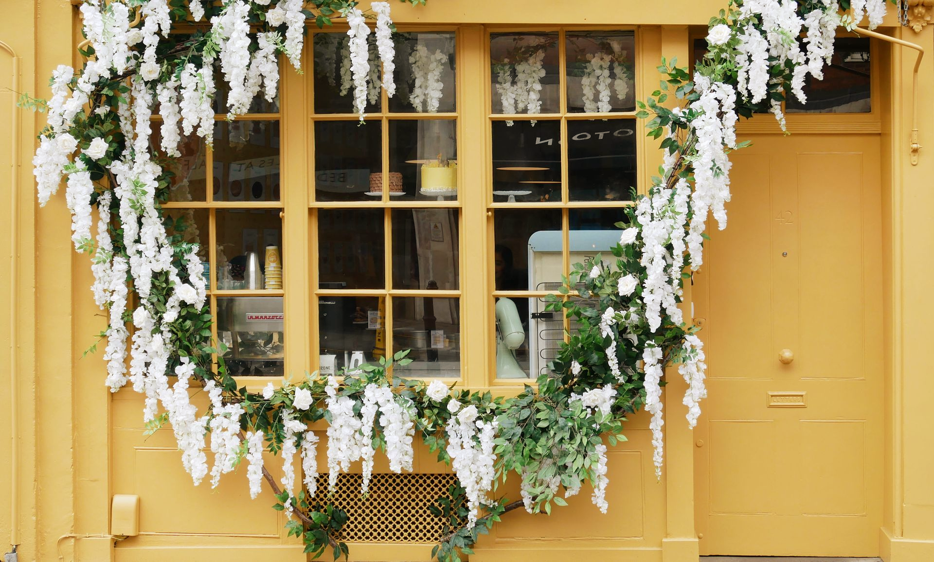 Une façade de magasin jaune moutarde ornée d'une guirlande luxuriante de glycines blanches encadrant une fenêtre et une porte.
