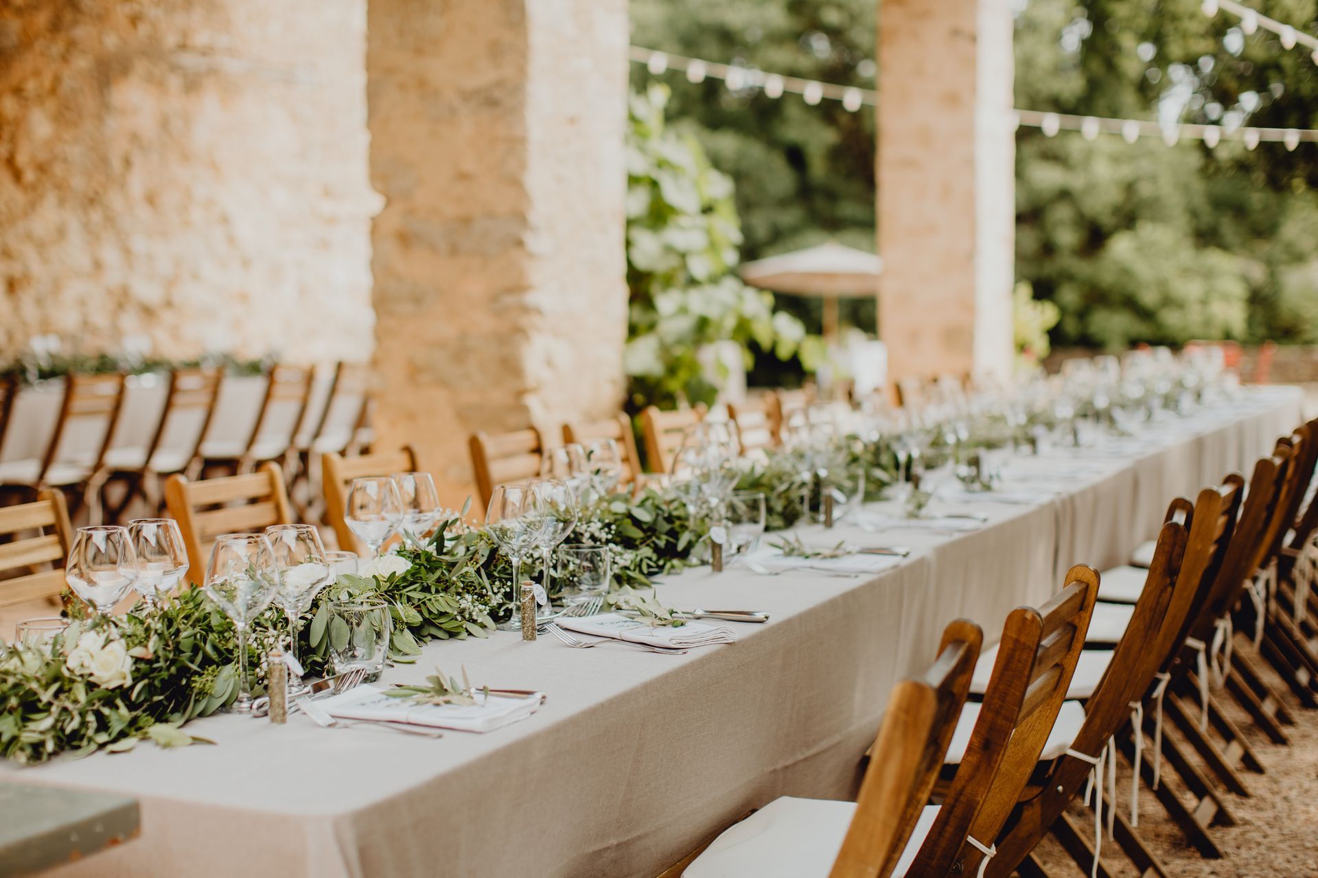 Une longue et élégante table de banquet dressée pour un mariage, avec des chemins de table végétaux, des verres à vin et des chaises sur une terrasse en pierre.