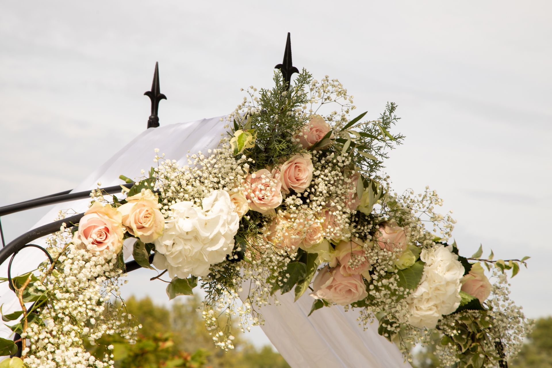 Une arche de mariage en fer forgé, ornée d'hortensias blancs, de roses rose pâle et de gypsophile, se détachant sur un ciel clair.