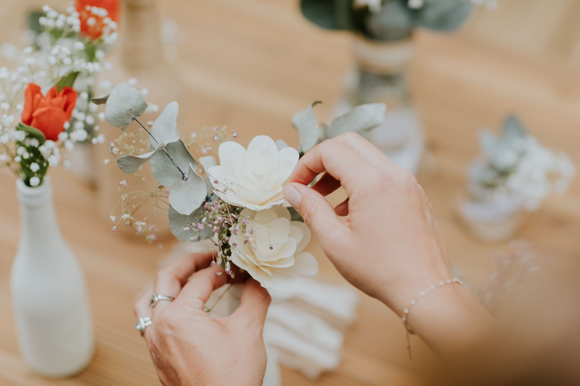 Des mains disposent sur une table une délicate boutonnière florale composée de fleurs de bois blanc, de feuilles d'eucalyptus et de gypsophile.