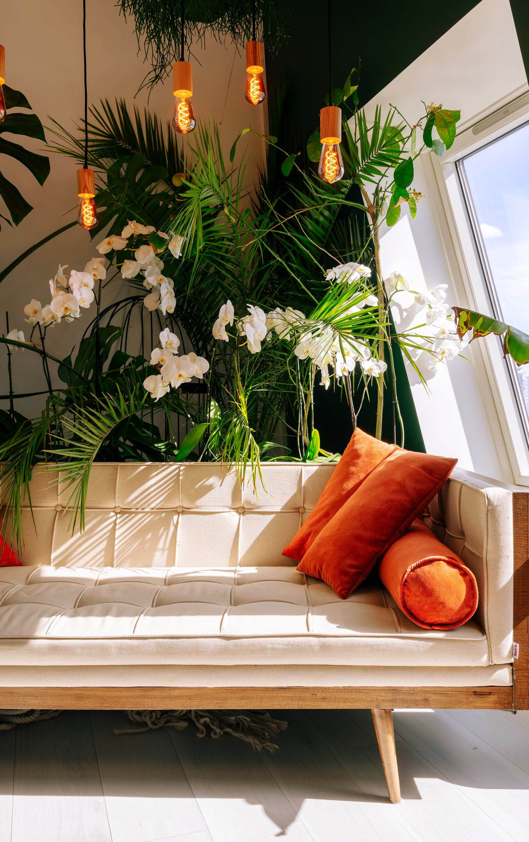 Dans une pièce lumineuse et ensoleillée, une méridienne aux coussins orange est installée sous une végétation luxuriante et des ampoules suspendues.