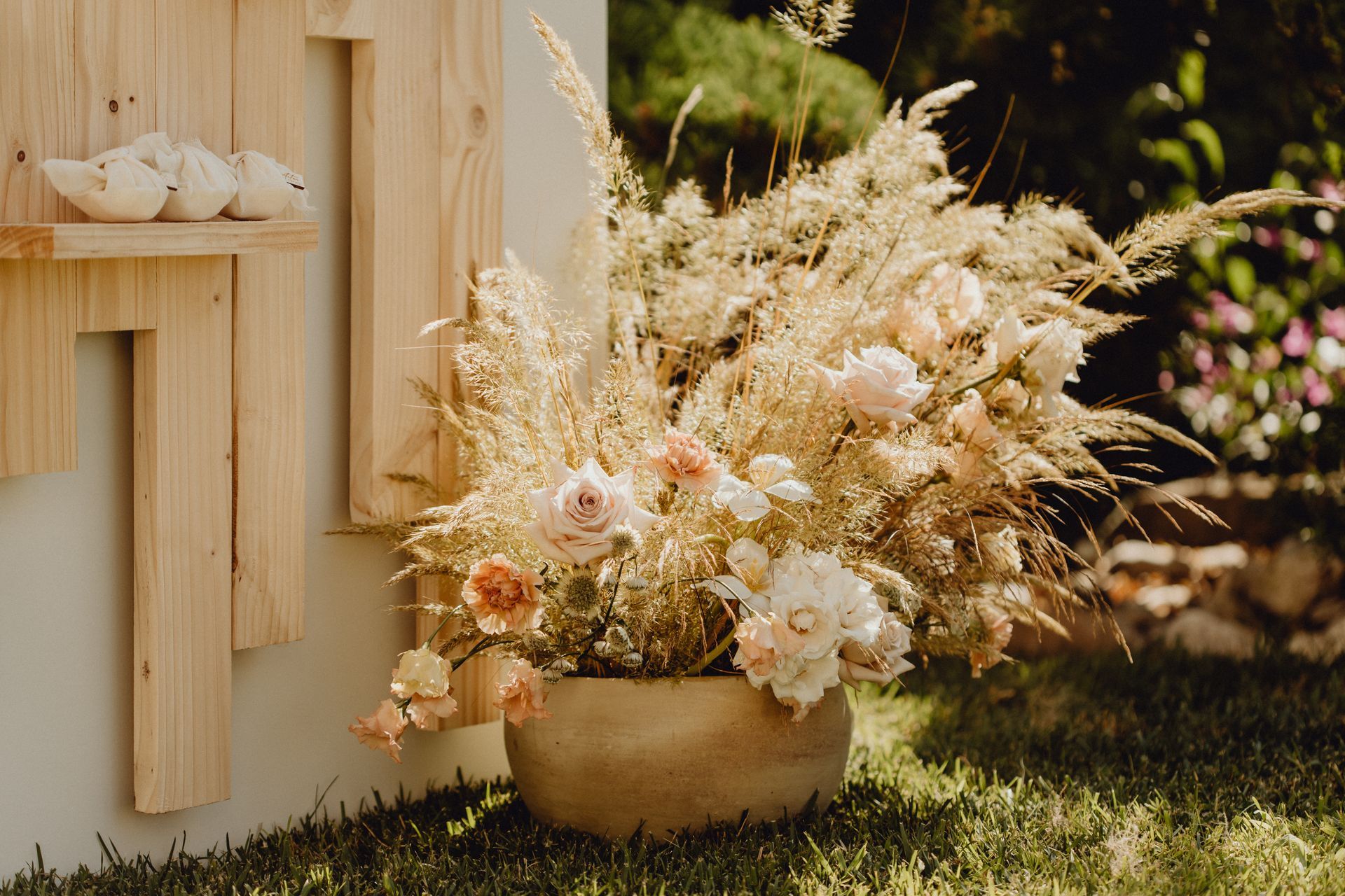 Une composition florale d'herbes séchées et de roses aux tons doux dans un bol en pierre, placée à l'extérieur près d'une installation murale en bois.