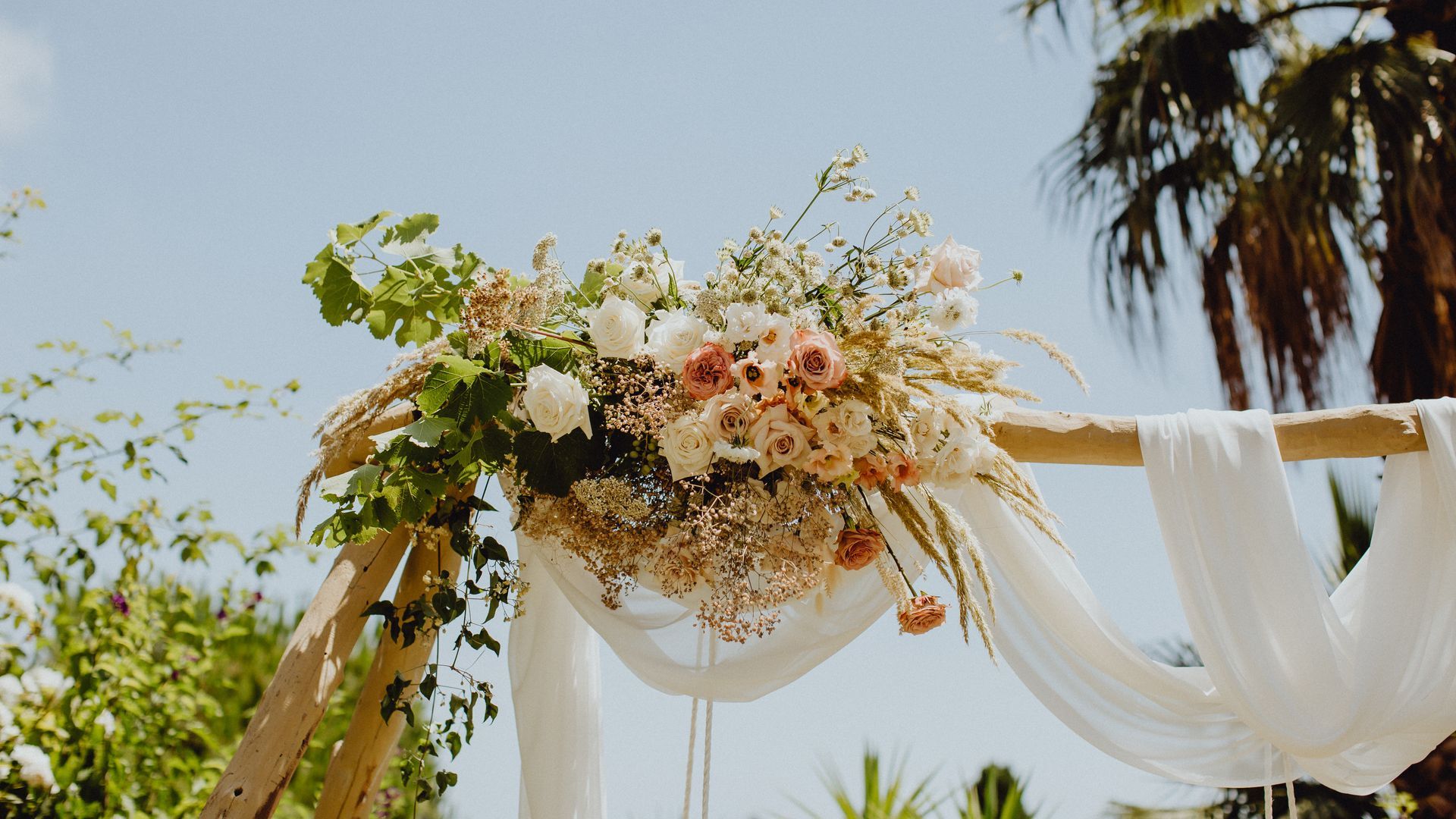 Une arche de mariage en bois ornée de tissu blanc, de verdure luxuriante et de compositions florales rose pâle et blanches.