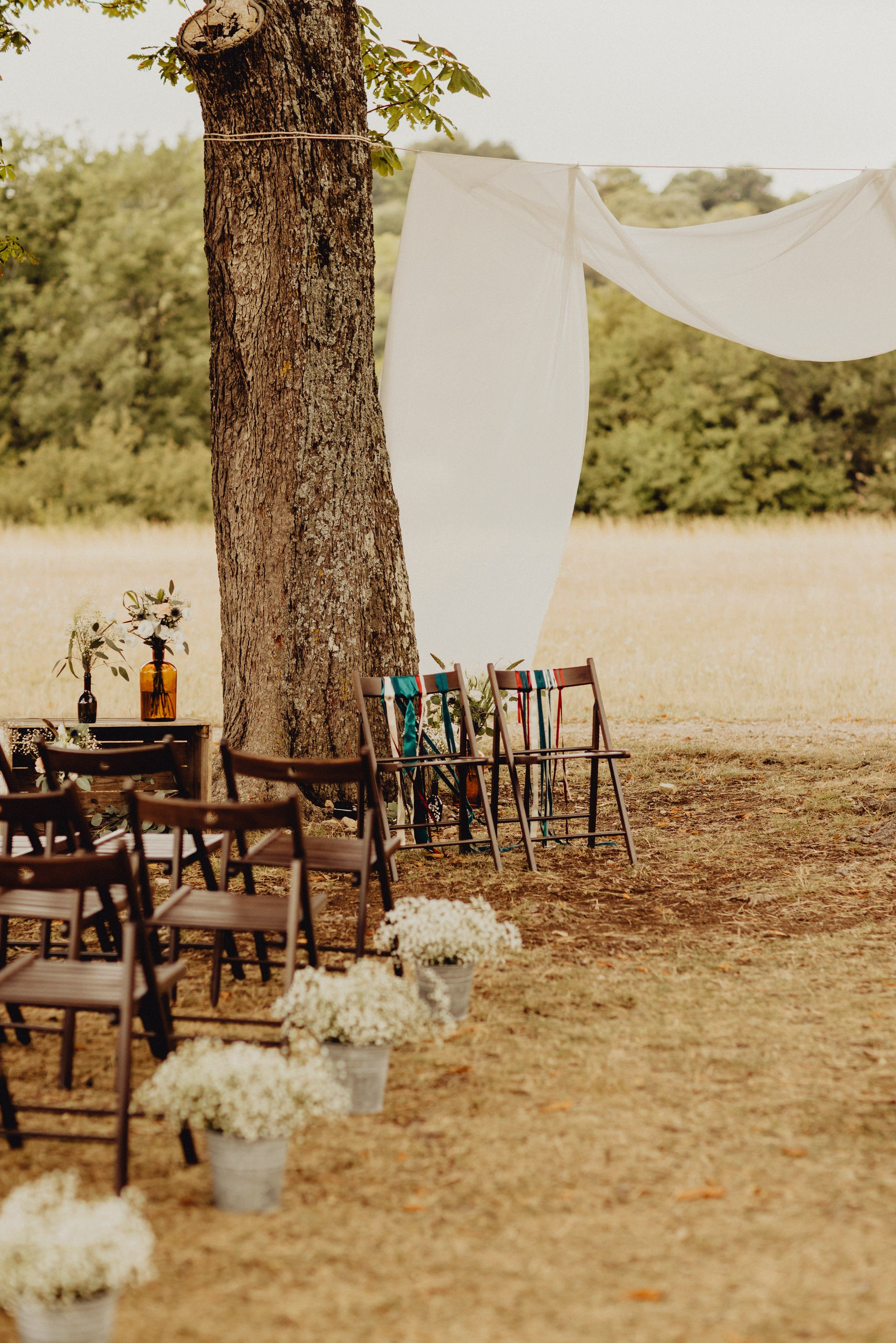 Un décor de mariage champêtre en plein air avec des chaises en bois, des touches florales blanches et un dais en tissu drapé depuis un grand arbre.