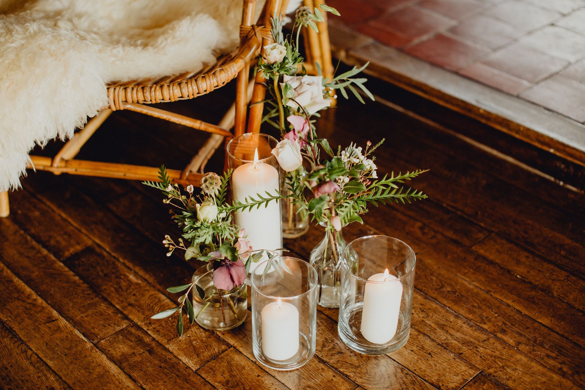 Des bougies blanches dans des cylindres de verre et de petits vases contenant de la verdure et des fleurs violettes sont disposés sur un parquet près d'une chaise en rotin.