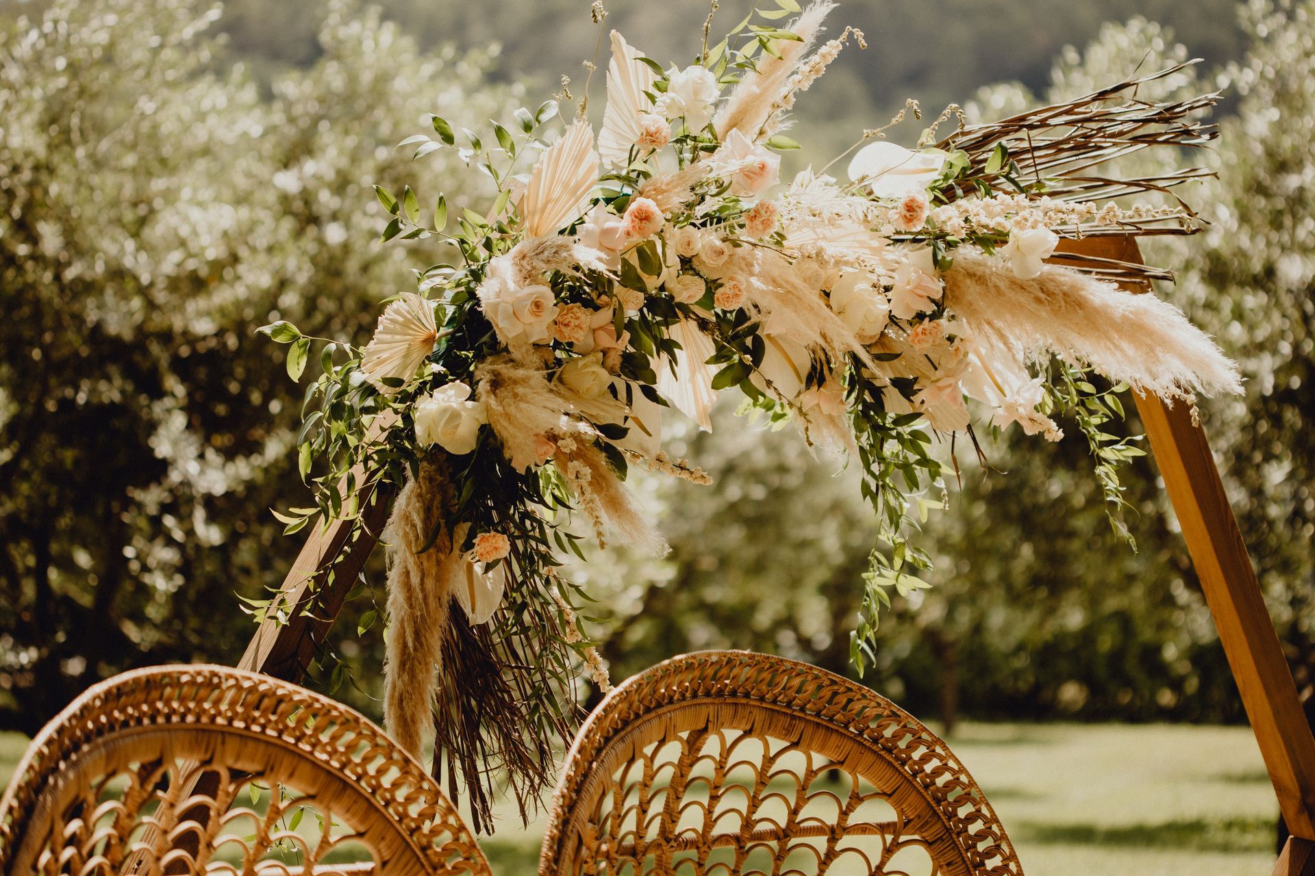 Une arche florale décorative composée d'herbes de la pampa et de roses rose pâle, placée derrière deux chaises en osier tressé dans un jardin.