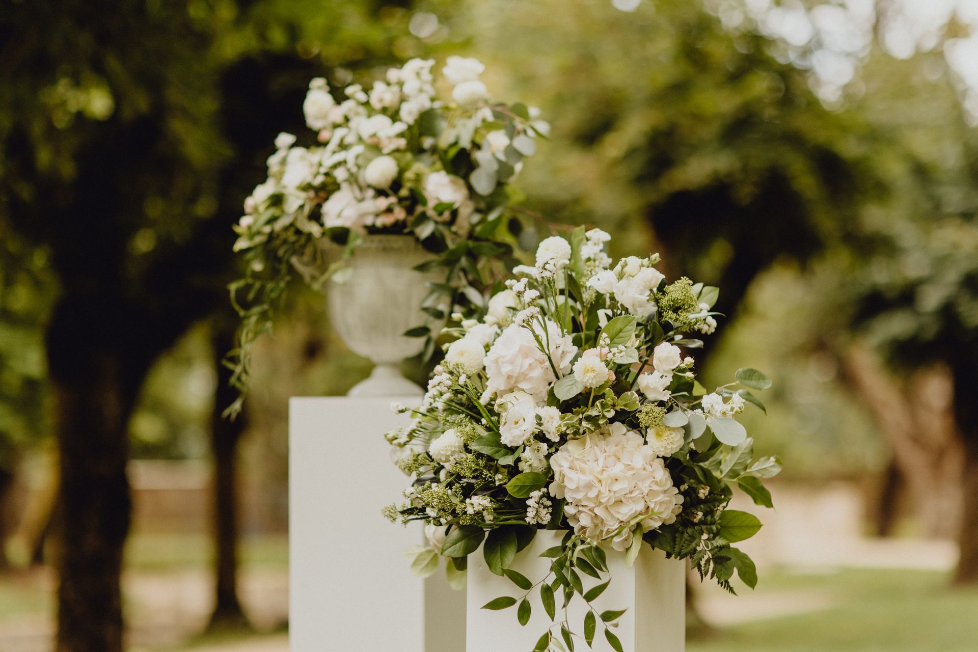 Deux compositions florales de fleurs blanches et de verdure reposent sur des socles blancs dans un parc.