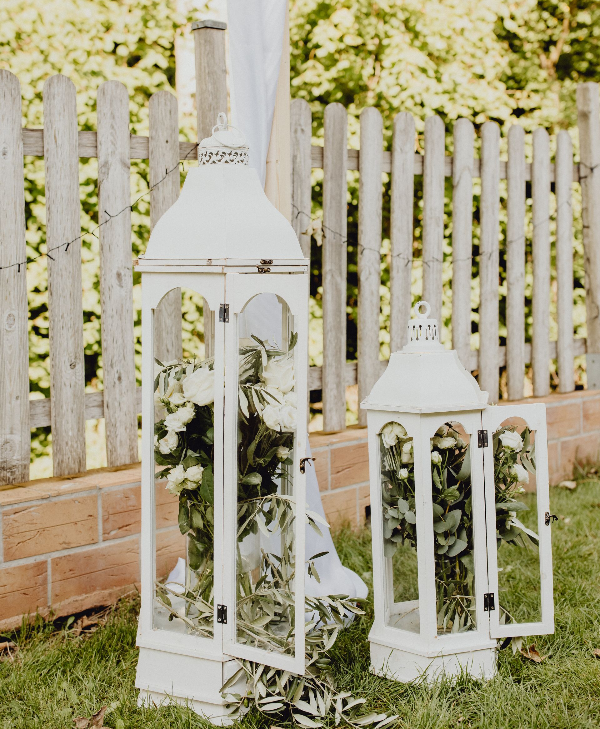 Deux lanternes décoratives blanches remplies de roses blanches et de verdure, posées sur l'herbe devant une clôture rustique en bois.