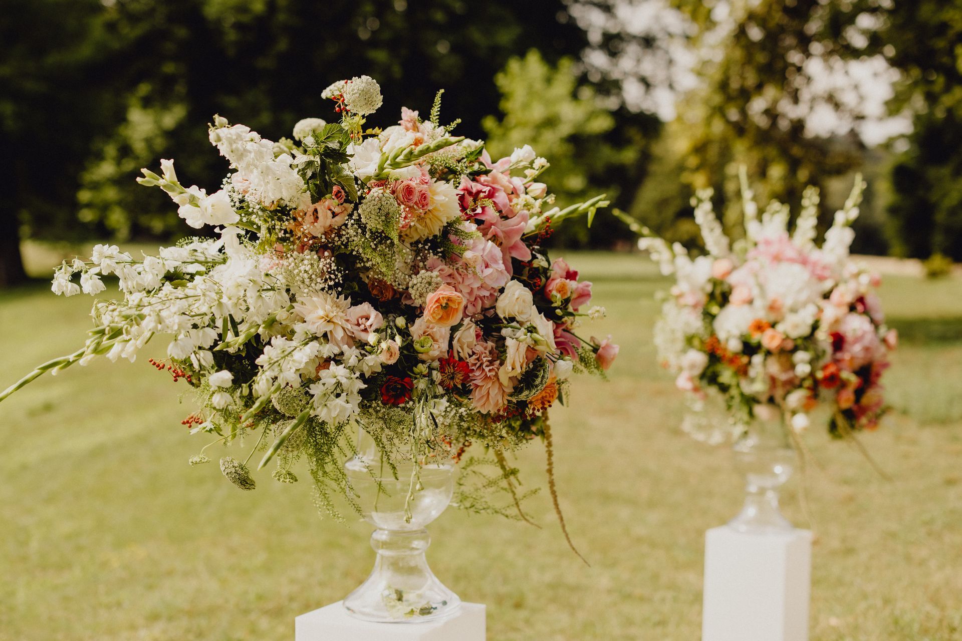 Deux grandes compositions florales aux couleurs éclatantes, présentées dans des vases en verre, reposent sur des socles blancs au milieu d'un champ herbeux.