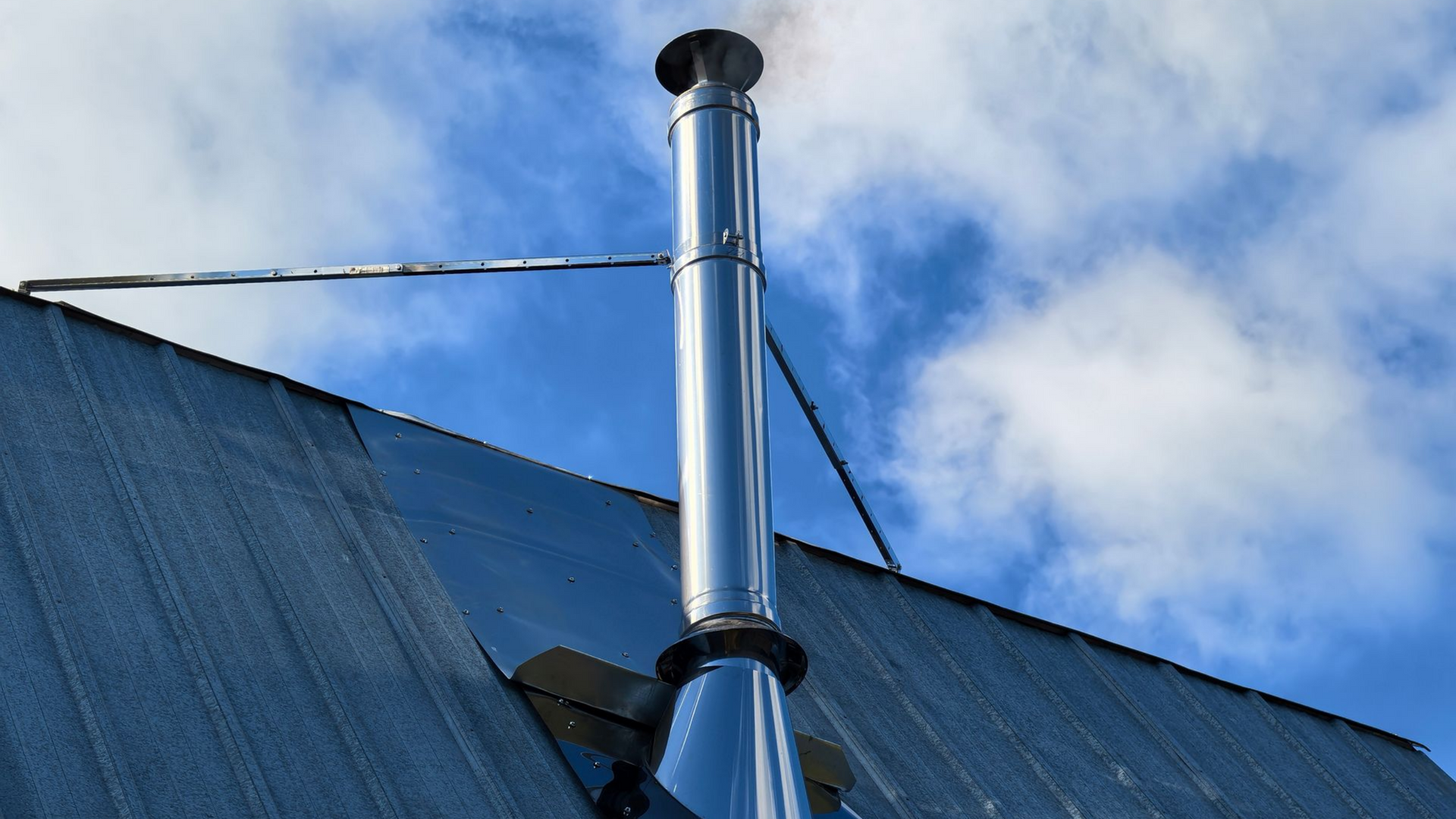 Cheminée en métal brillant sur un toit en tôle ondulée bleue, fumée s'élevant sur un ciel nuageux.