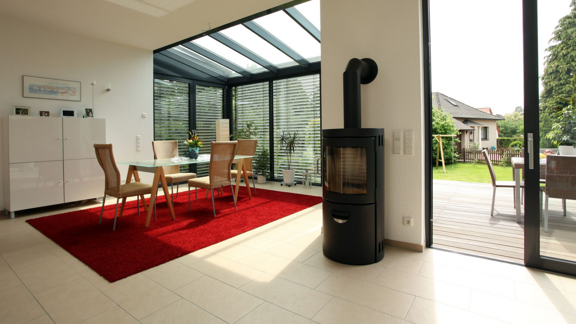 Salle à manger avec toit en verre, tapis rouge, cheminée et porte ouverte sur une cour arrière.