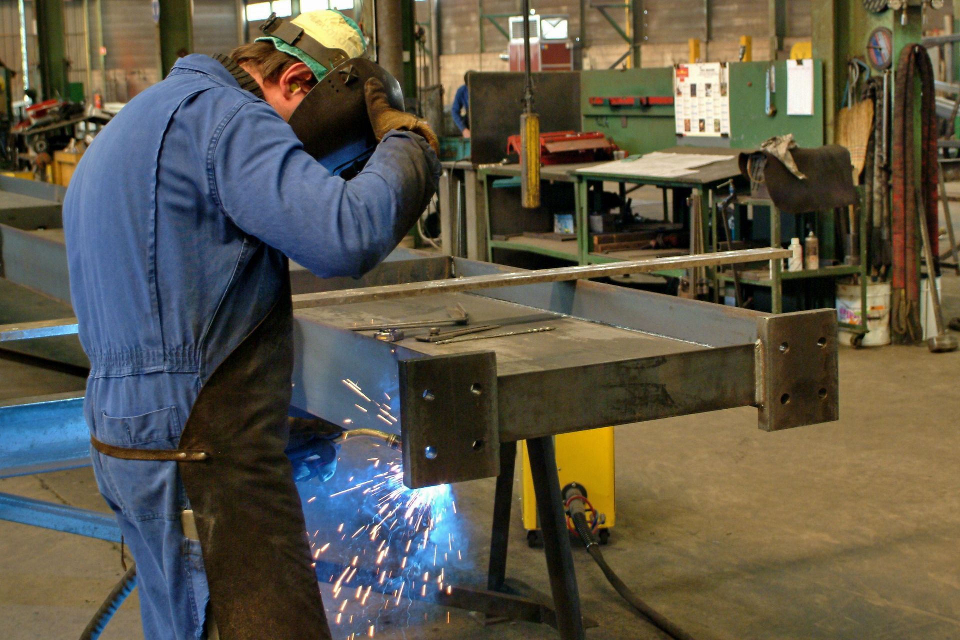 Soudeur en salopette bleue en train de souder une structure métallique, des étincelles jaillissent, intérieur d'usine.