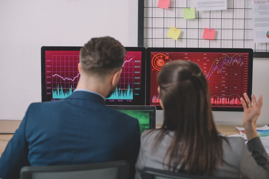 Dos personas mirando gráficos bursátiles en rojo en monitores de computadora; entorno de oficina.