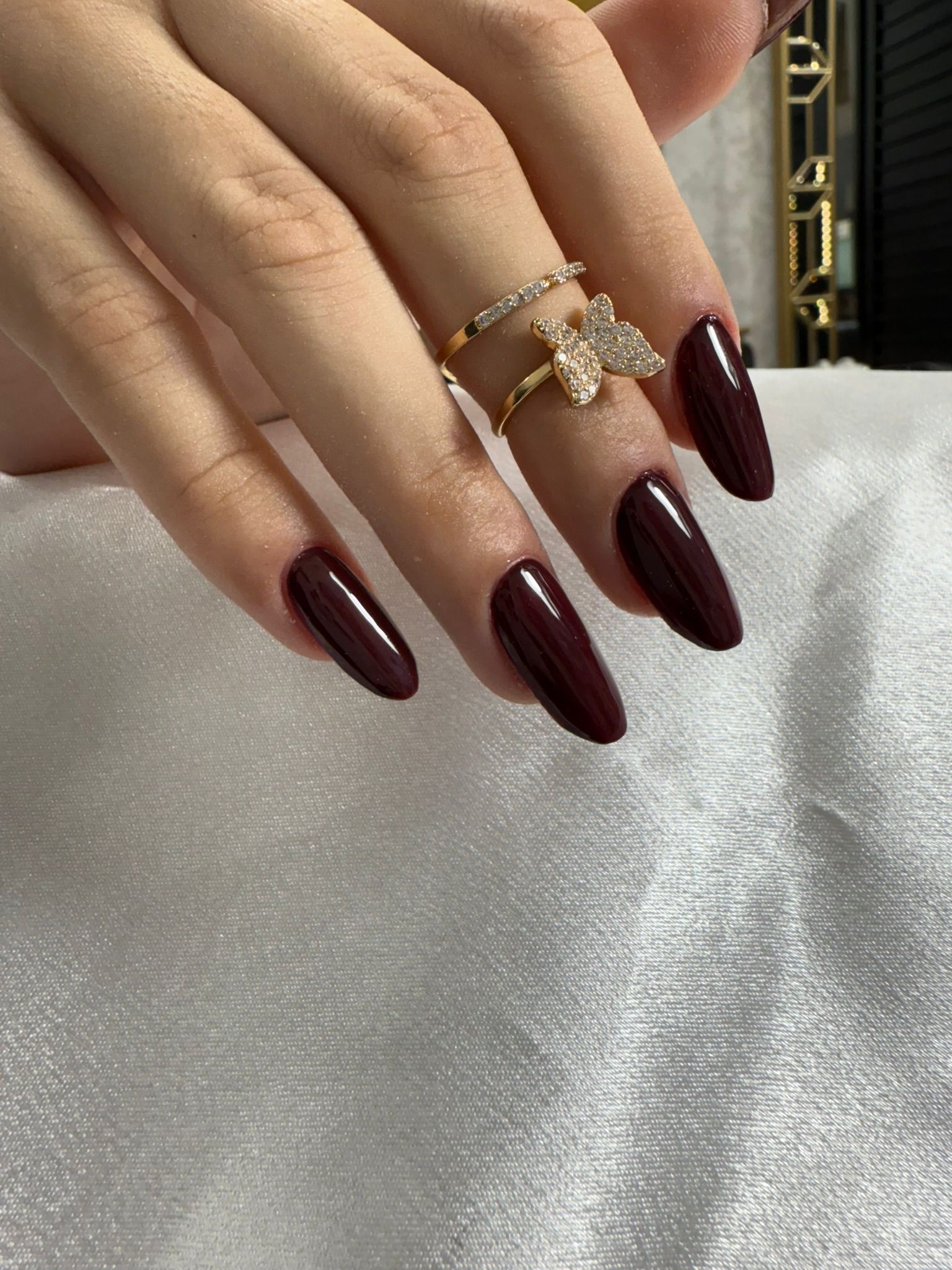 Mano con uñas largas de color rojo oscuro y dos anillos de oro sobre fondo blanco.