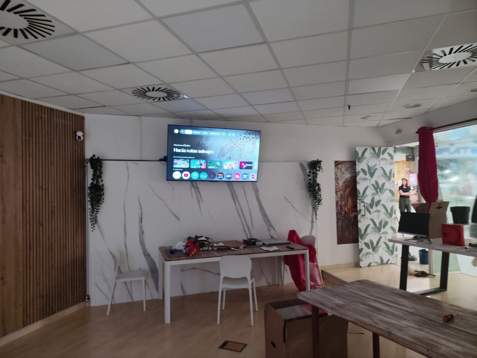 Salón moderno con televisor montado en la pared, mesas y sillas blancas, paneles de madera y plantas colgantes
