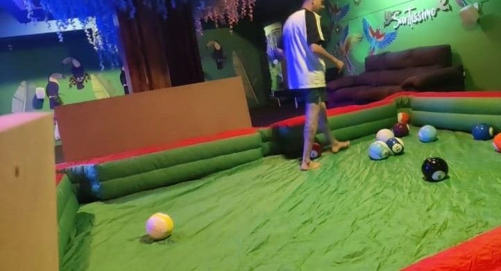Zona de juegos interior con suelo acolchado verde, pelotas esparcidas y un niño de pie junto a un mural en la pared.
