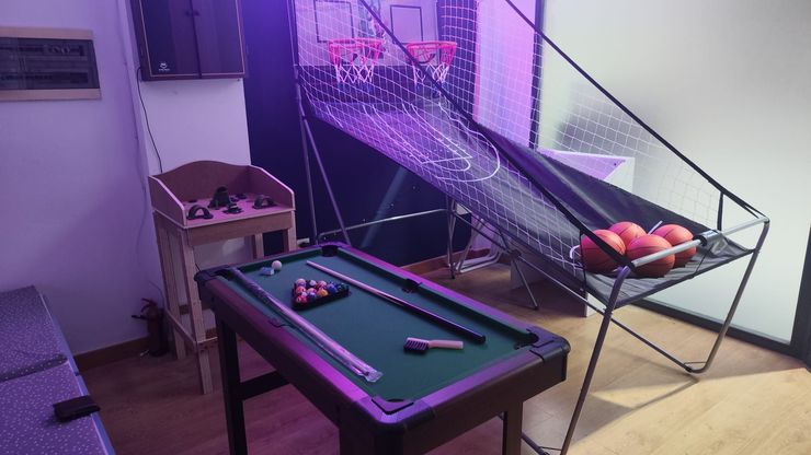 Sala de juegos estilo arcade con mesa de billar, mesa de hockey de aire y una canasta de baloncesto sobre un soporte para pelotas