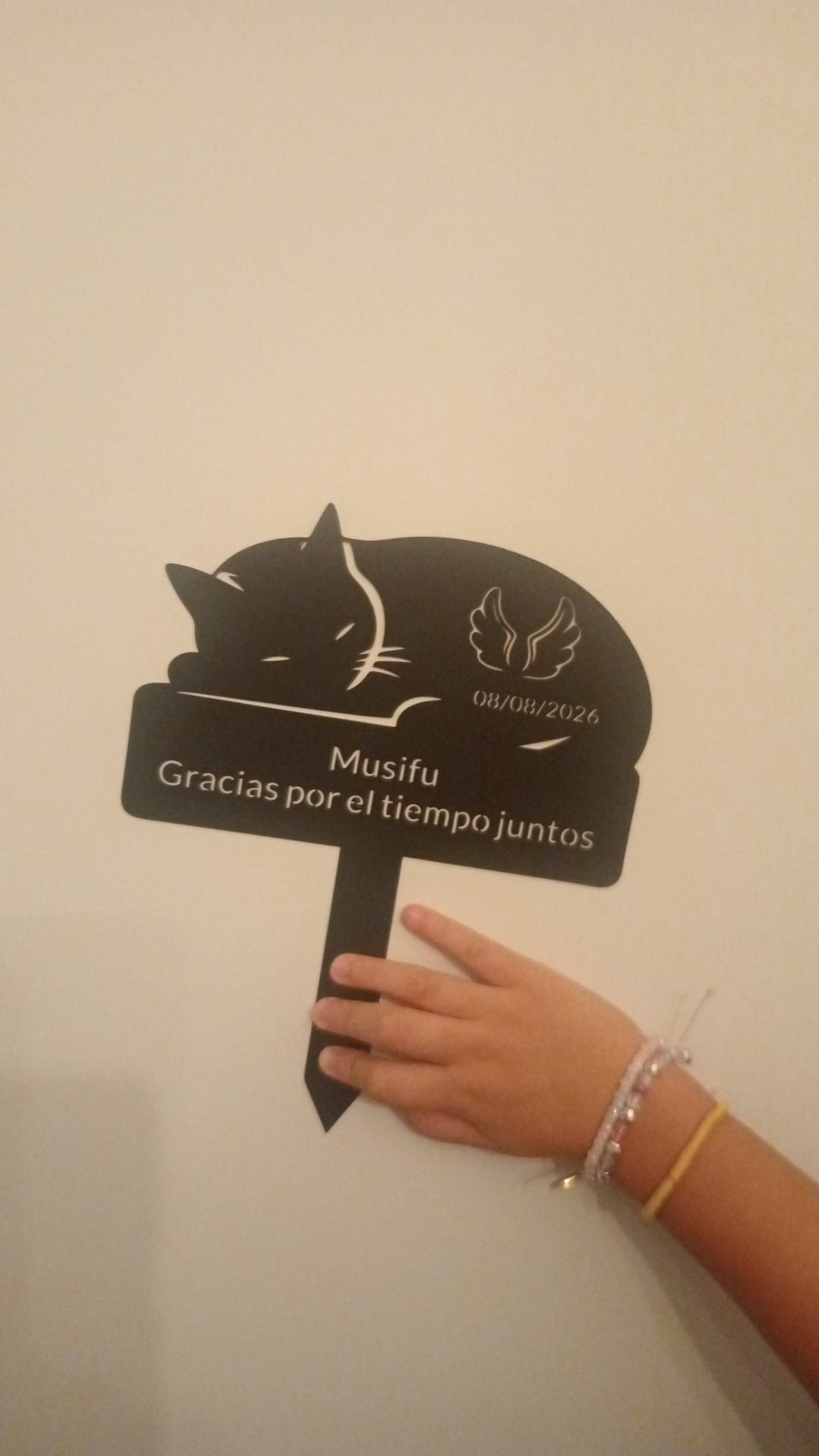 Una mano sostiene un cartel negro con forma de gato y texto blanco frente a una pared beige.