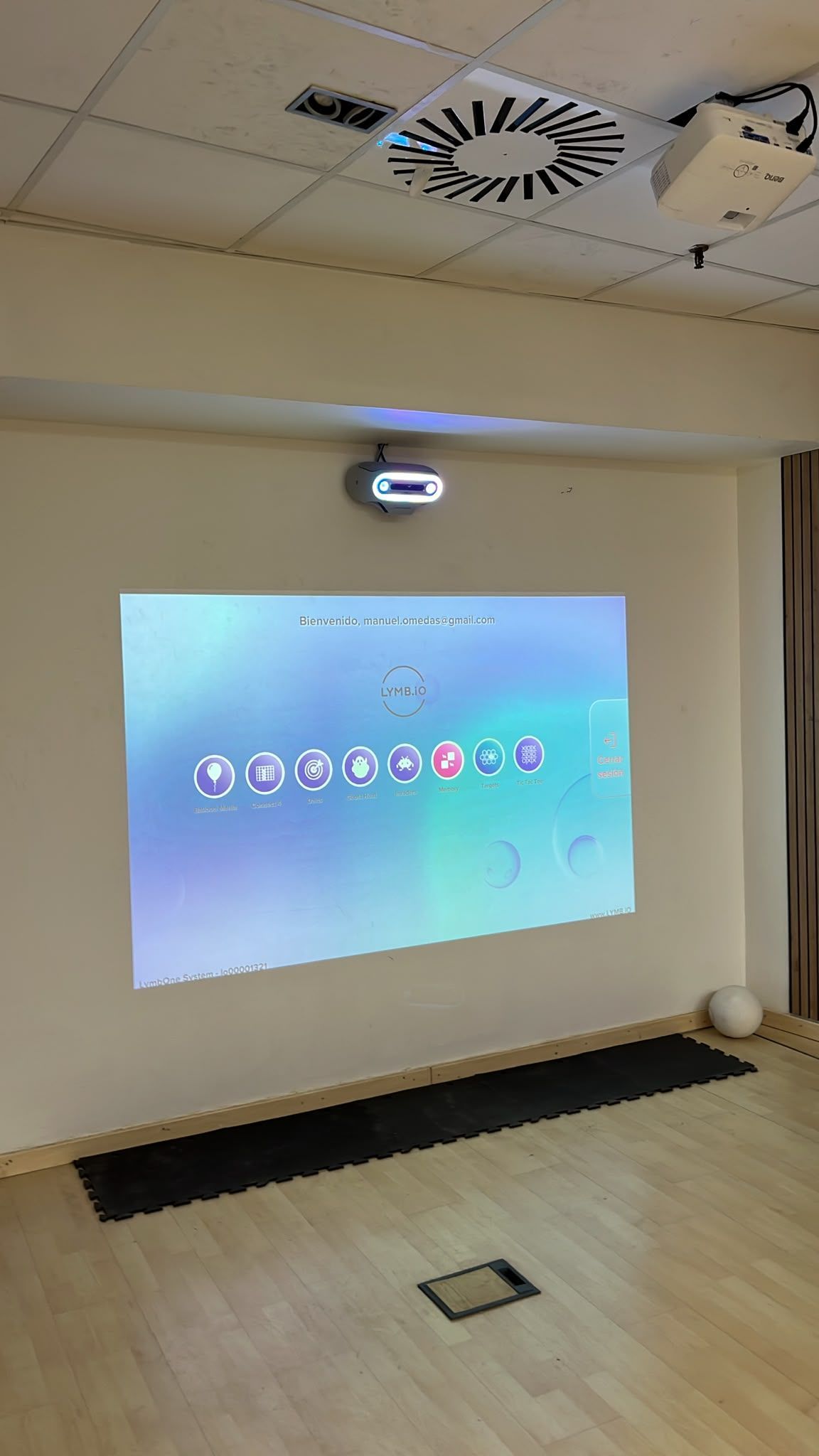 Sala vacía con un proyector que muestra un escritorio en la pared, suelo de madera clara y rejillas de ventilación visibles en el techo
