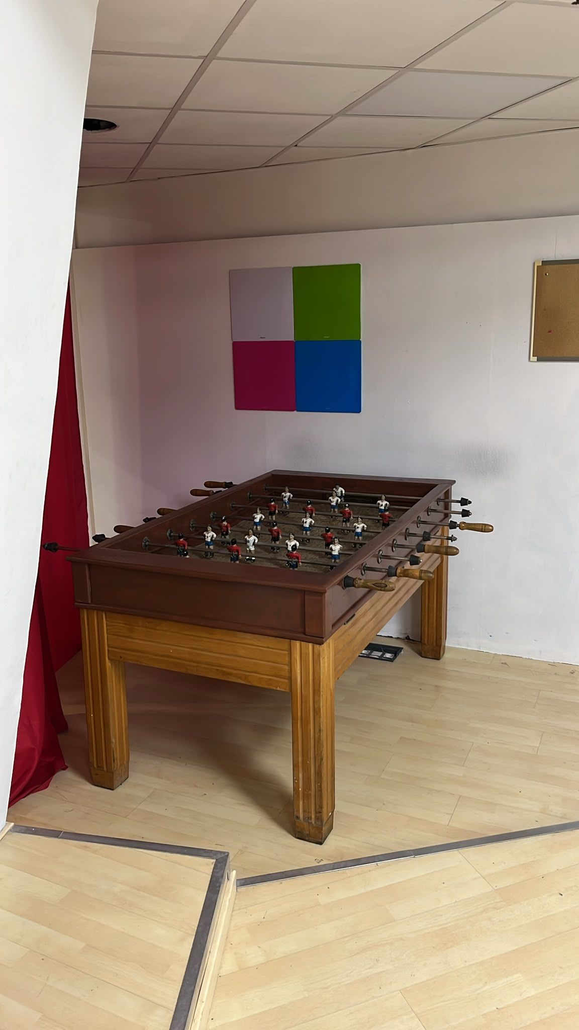 Futbolín en una habitación pequeña, con un cuadro cuadrado de colores en la pared y suelo de madera