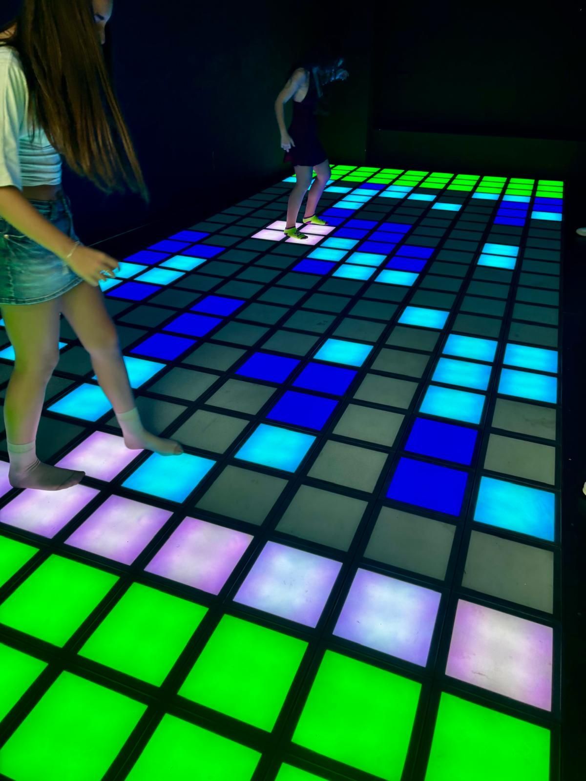 Personas bailando sobre un suelo de baldosas luminosas azules, verdes y moradas en una habitación oscura